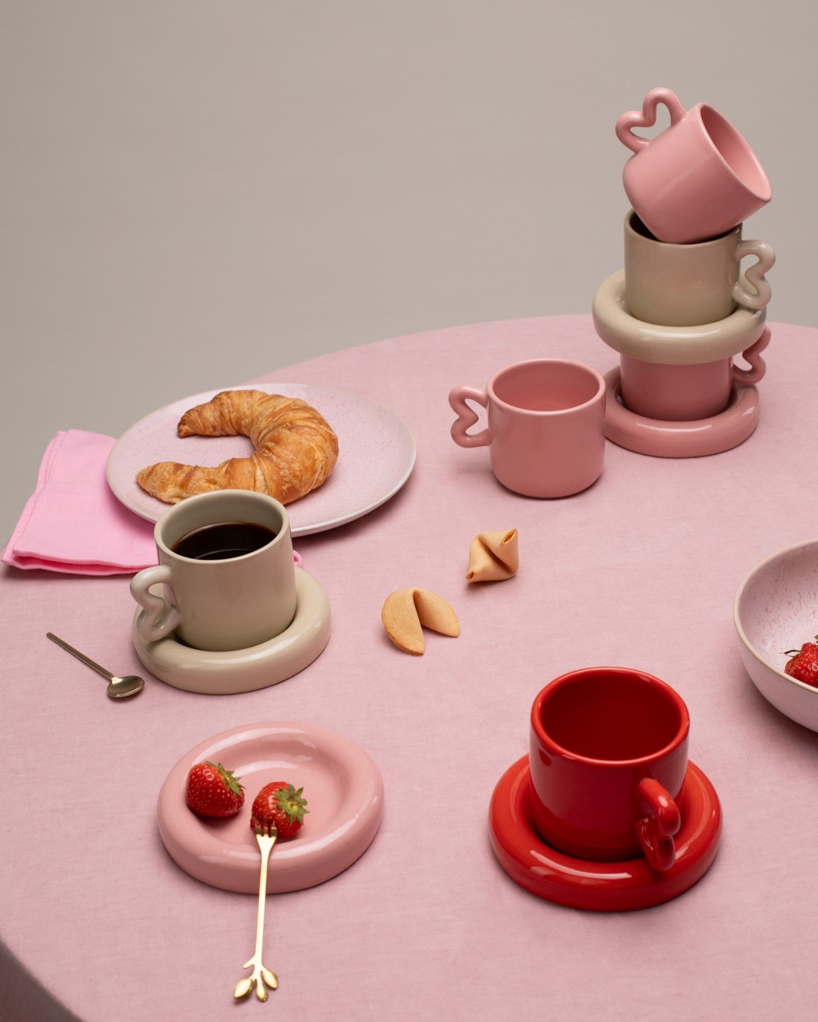 Verschiedene farbige Tassen, Croissant und Erdbeeren auf einem rosa Tisch mit elegantem Geschirr und Besteck.