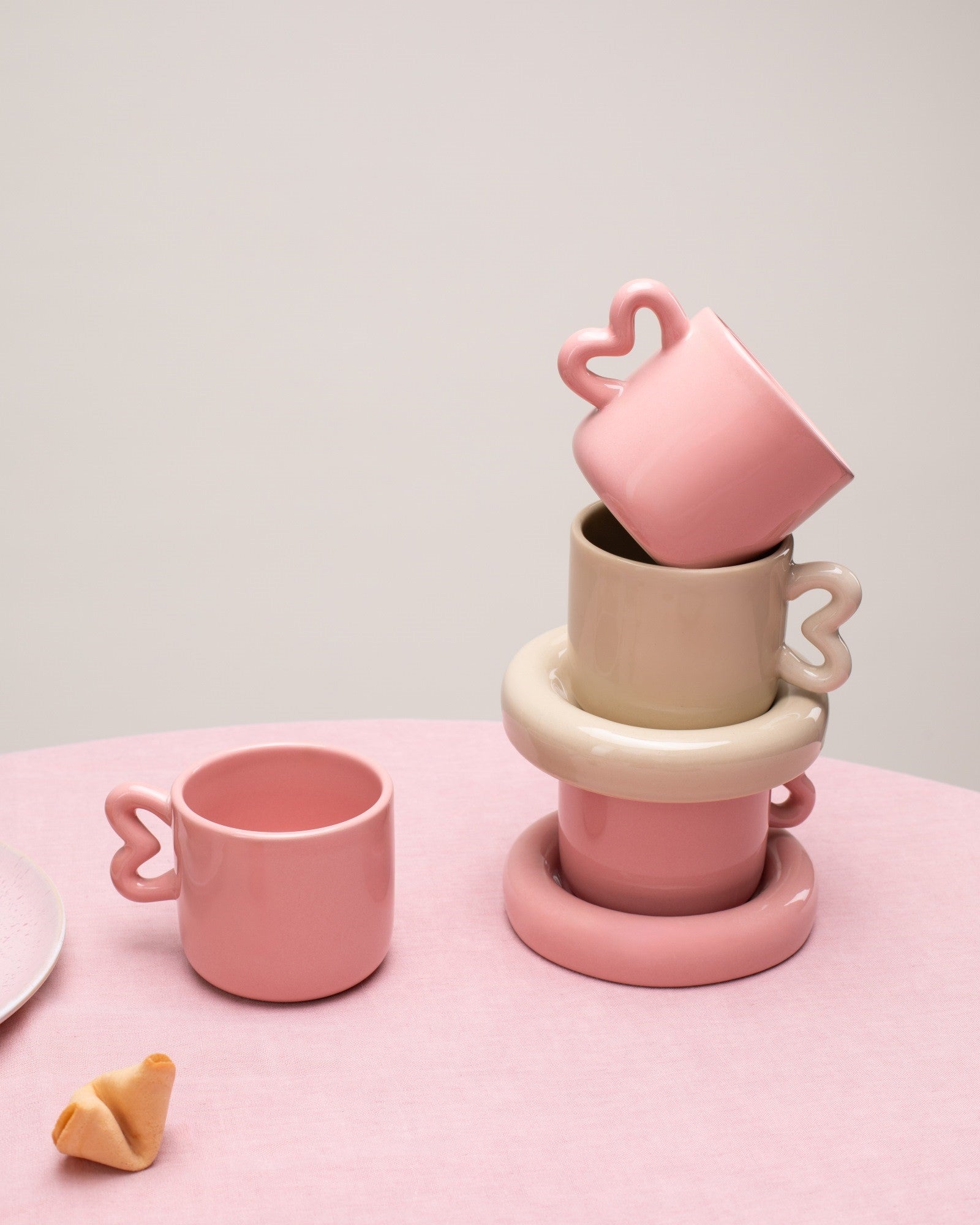 Rosafarbene und beigefarbene Tassen mit Herzgriffen sind auf einem rosa Tisch gestapelt; daneben liegt ein Glückskeks.