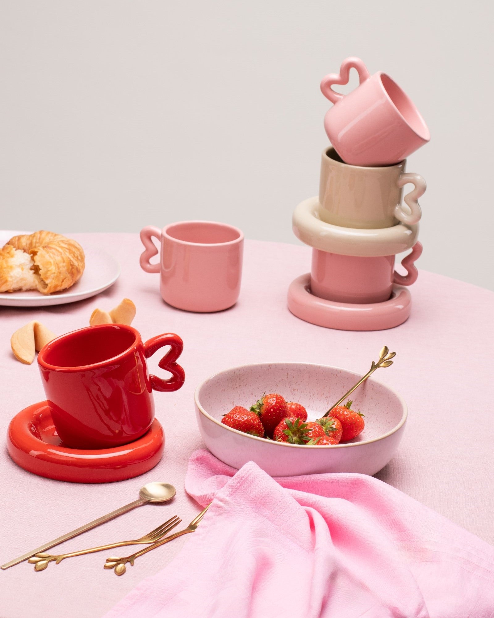 Rosafarbener Tisch mit stapelbaren Tassen, Croissant auf einem Teller, Keramikschale mit Erdbeeren und goldenem Besteck.
