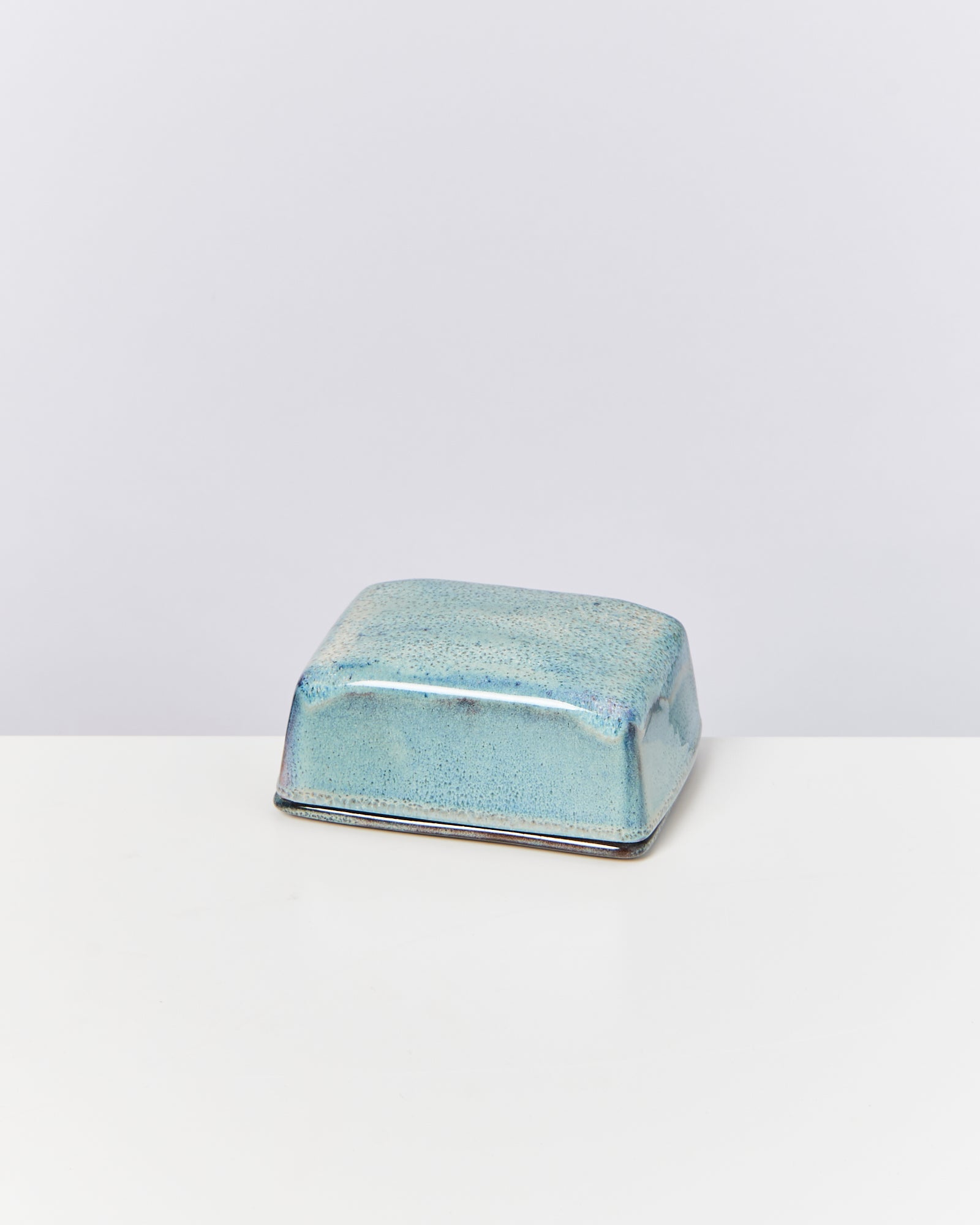 Ein quadratischer, türkisfarbener Keramikbehälter mit glänzender Glasur steht auf einer weißen Oberfläche.