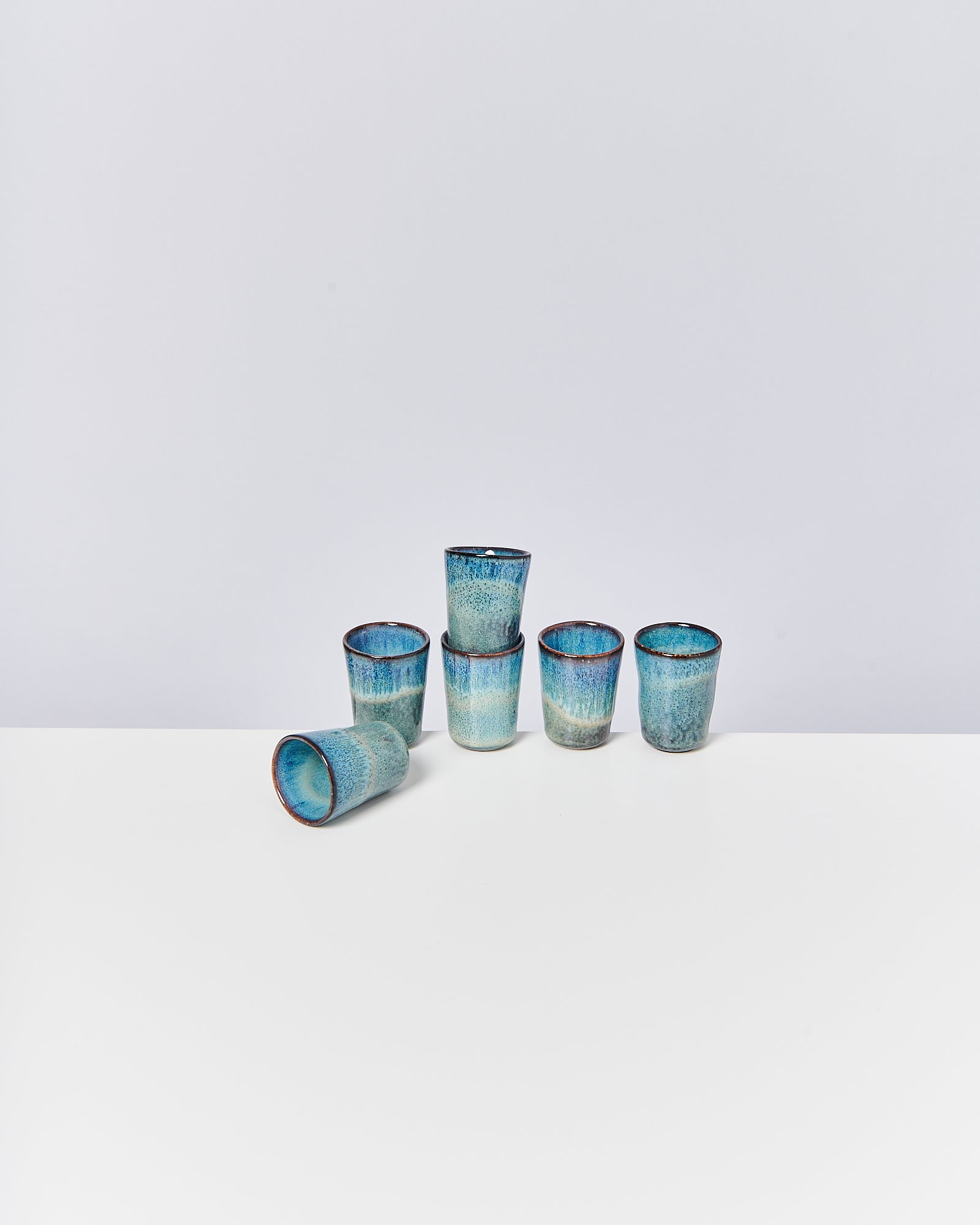 Sechs türkisfarbene Keramikbecher auf einem weißen Tisch, einer liegt seitlich.