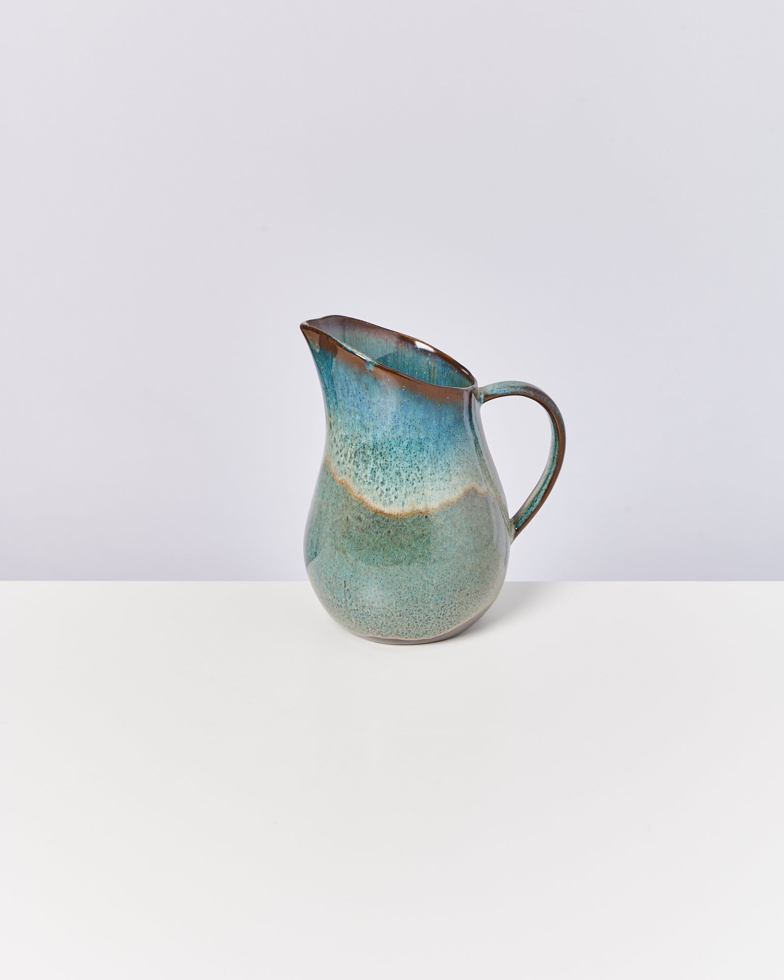 Grüner, keramischer Krug mit Henkel steht auf einem weißen Tisch vor neutralem Hintergrund.