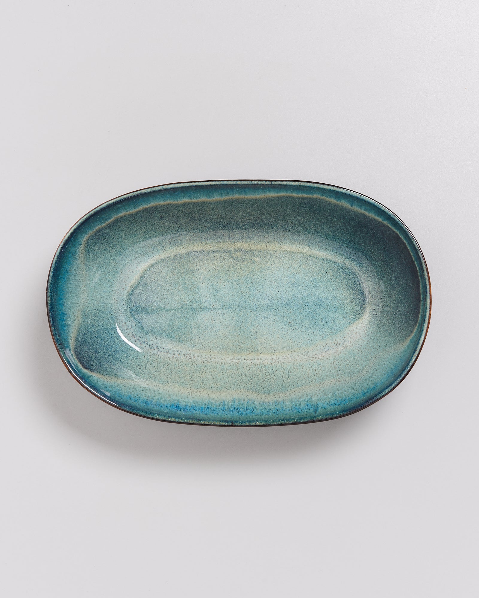Ovale, türkisfarbene Keramikplatte mit einem leicht welligen und glänzenden Finish auf weißem Hintergrund.