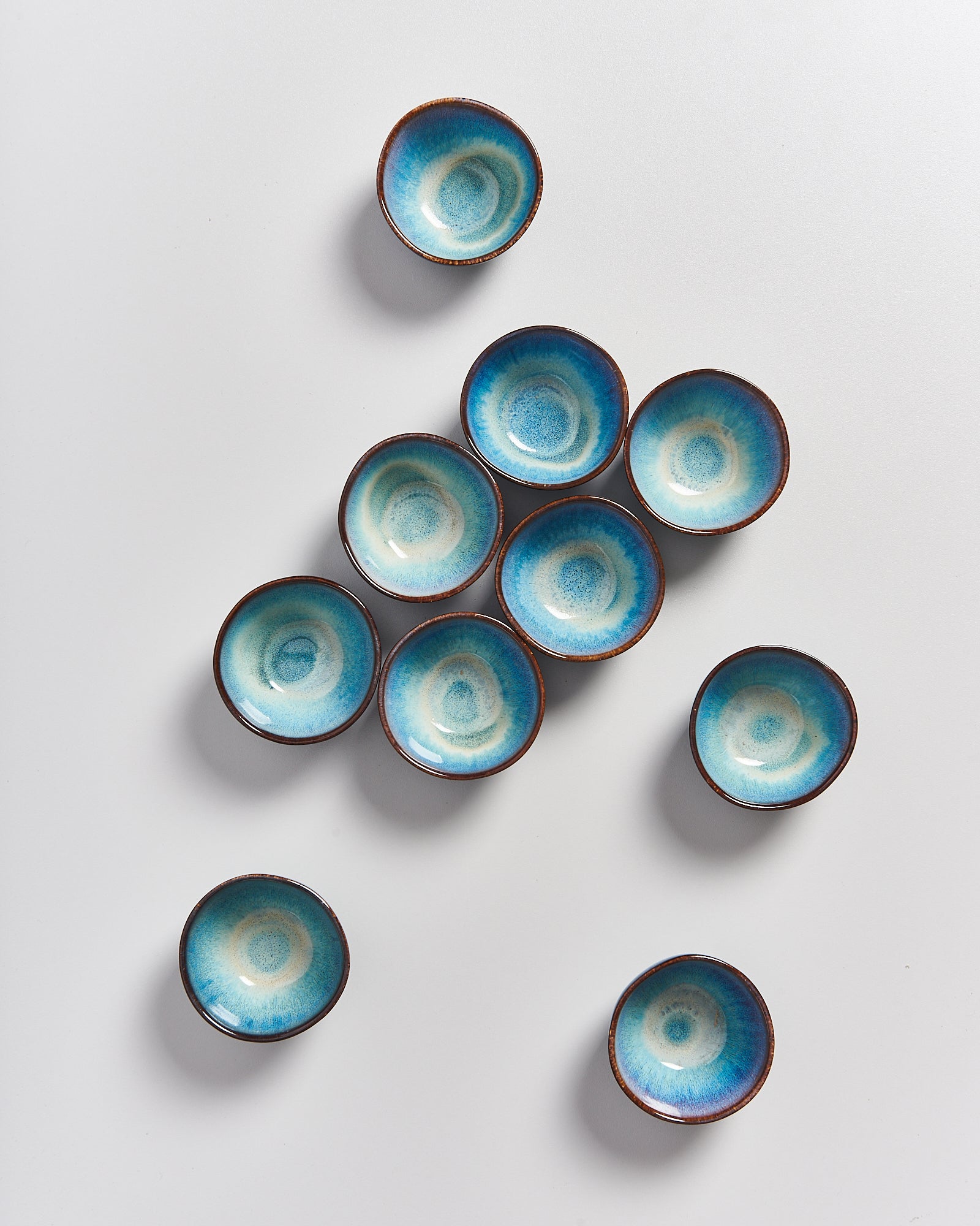 Zehn kleine, blaue Schalen mit braunem Rand liegen auf einem weißen Hintergrund verstreut.