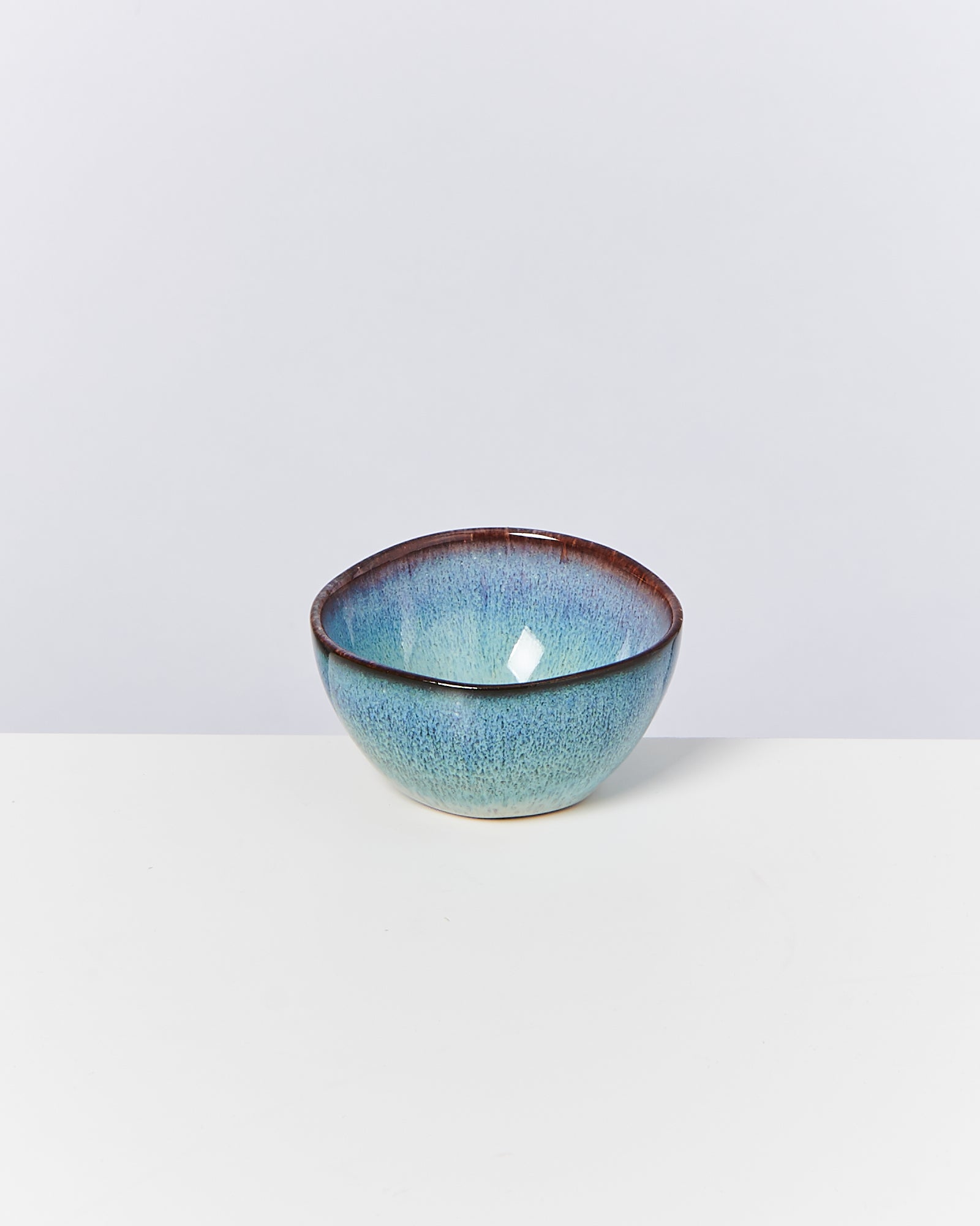 Eine kleine, blau glasierte Schüssel mit braunem Rand steht auf einem weißen Tisch vor einem grauen Hintergrund.