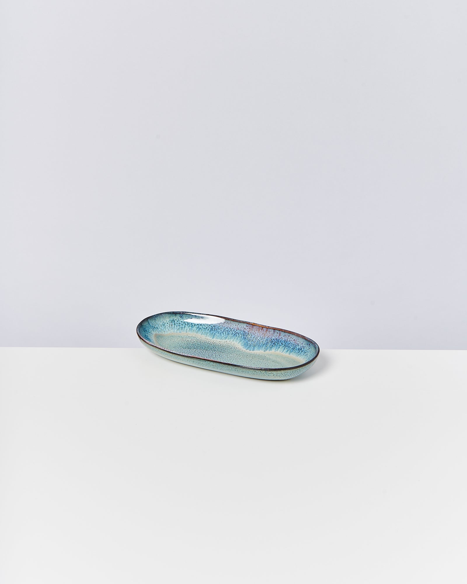 Langes, ovales Keramikschälchen in Blau- und Grüntönen auf einem weißen Tisch vor neutralem Hintergrund.