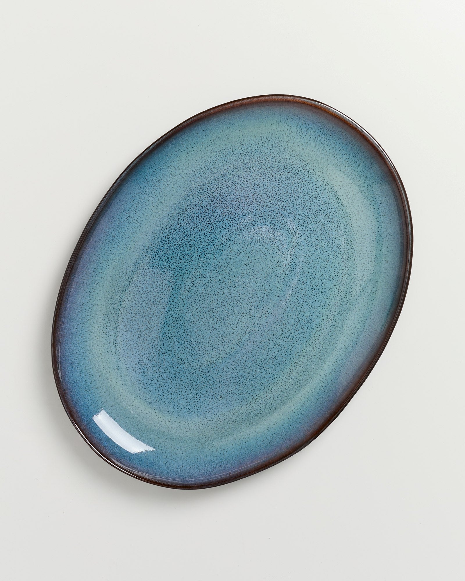 Ovale Keramikplatte mit blauer Glasur und dunkelbraunem Rand auf weißem Hintergrund.