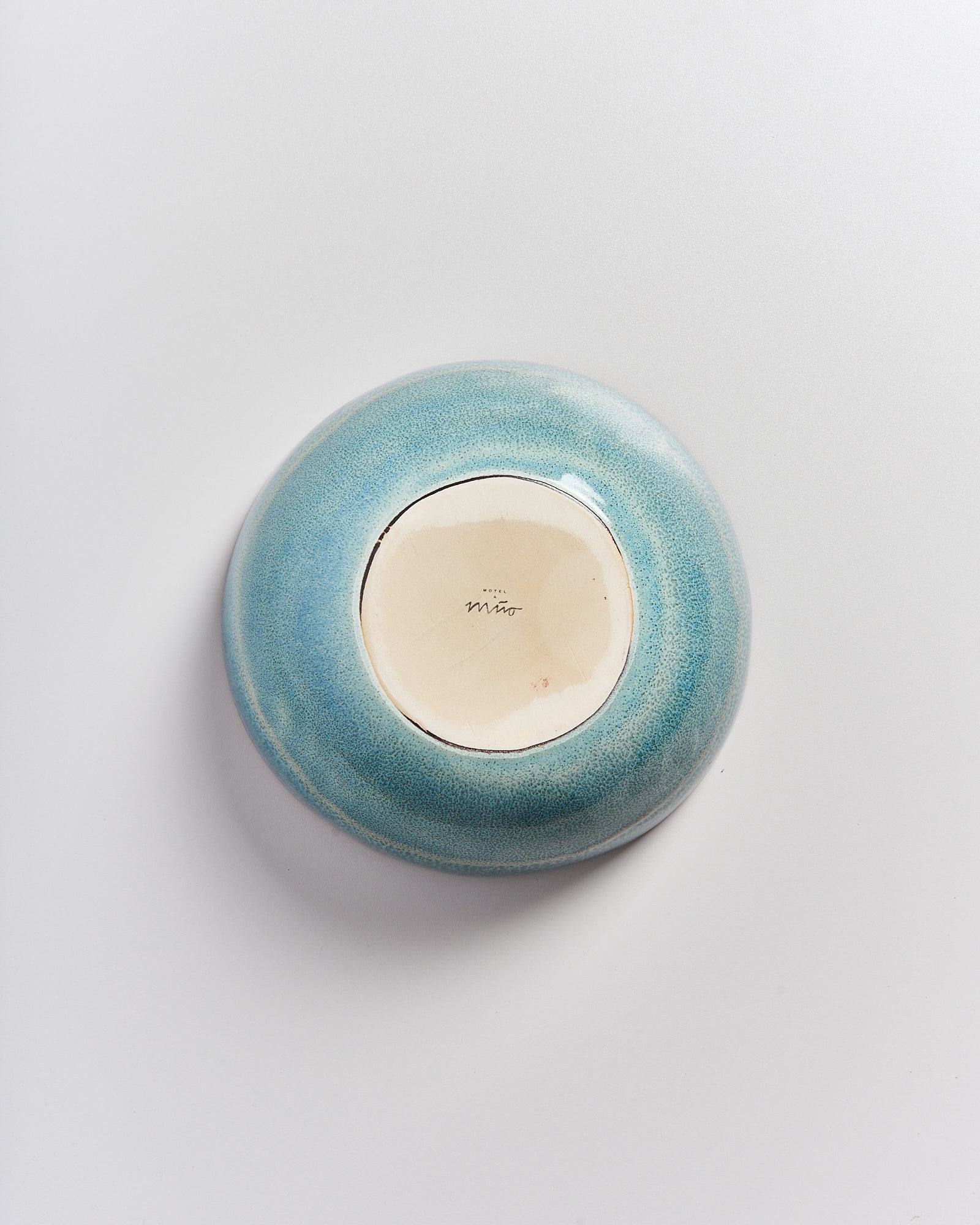 Die Unterseite einer blauen Keramikschale mit hellem Zentrum und einer unleserlichen Signatur auf weißem Hintergrund.