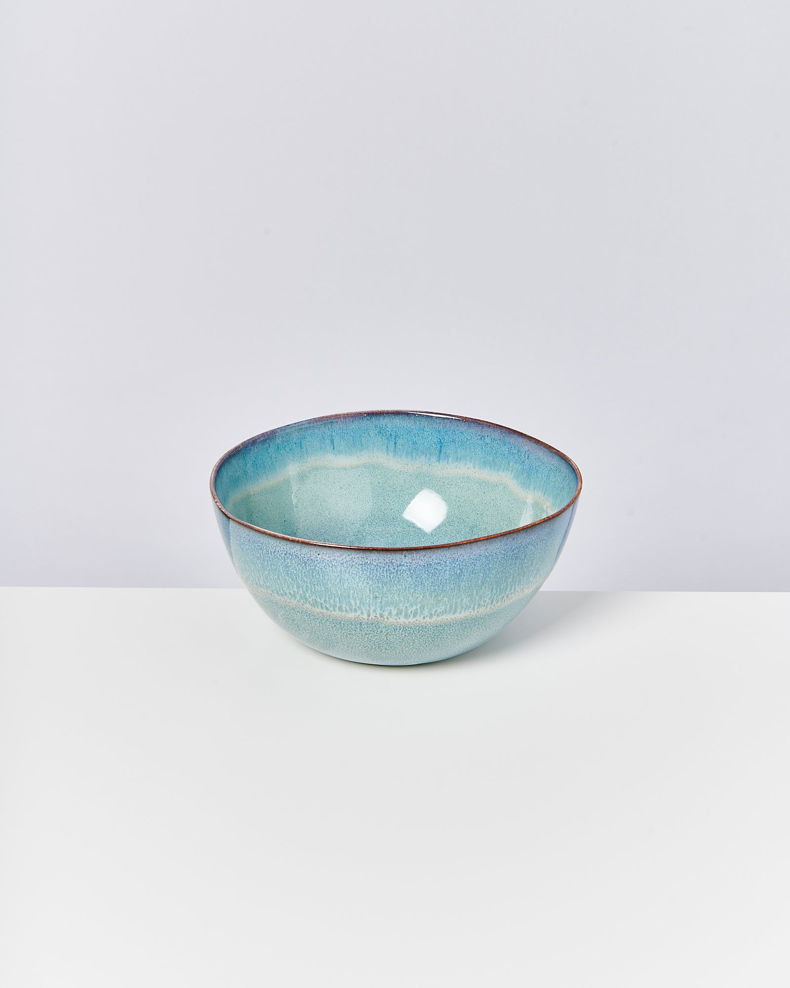 Türkisblaue, glasierte Keramikschale mit kunstvollen Farbverläufen auf einem weißen Tisch vor grauem Hintergrund.