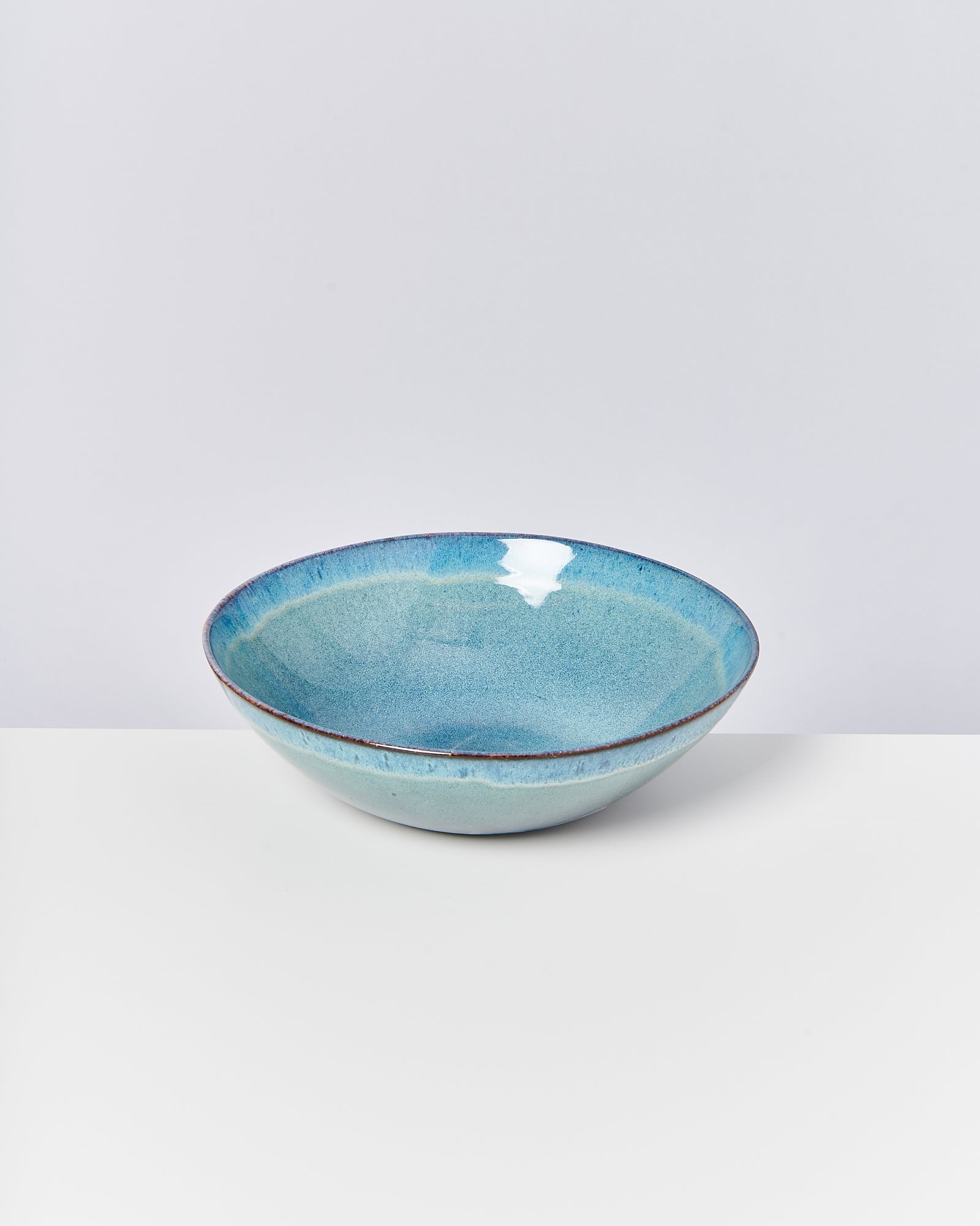 Eine blau-grüne Keramikschale steht auf einem weißen Untergrund vor einem hellgrauen Hintergrund.