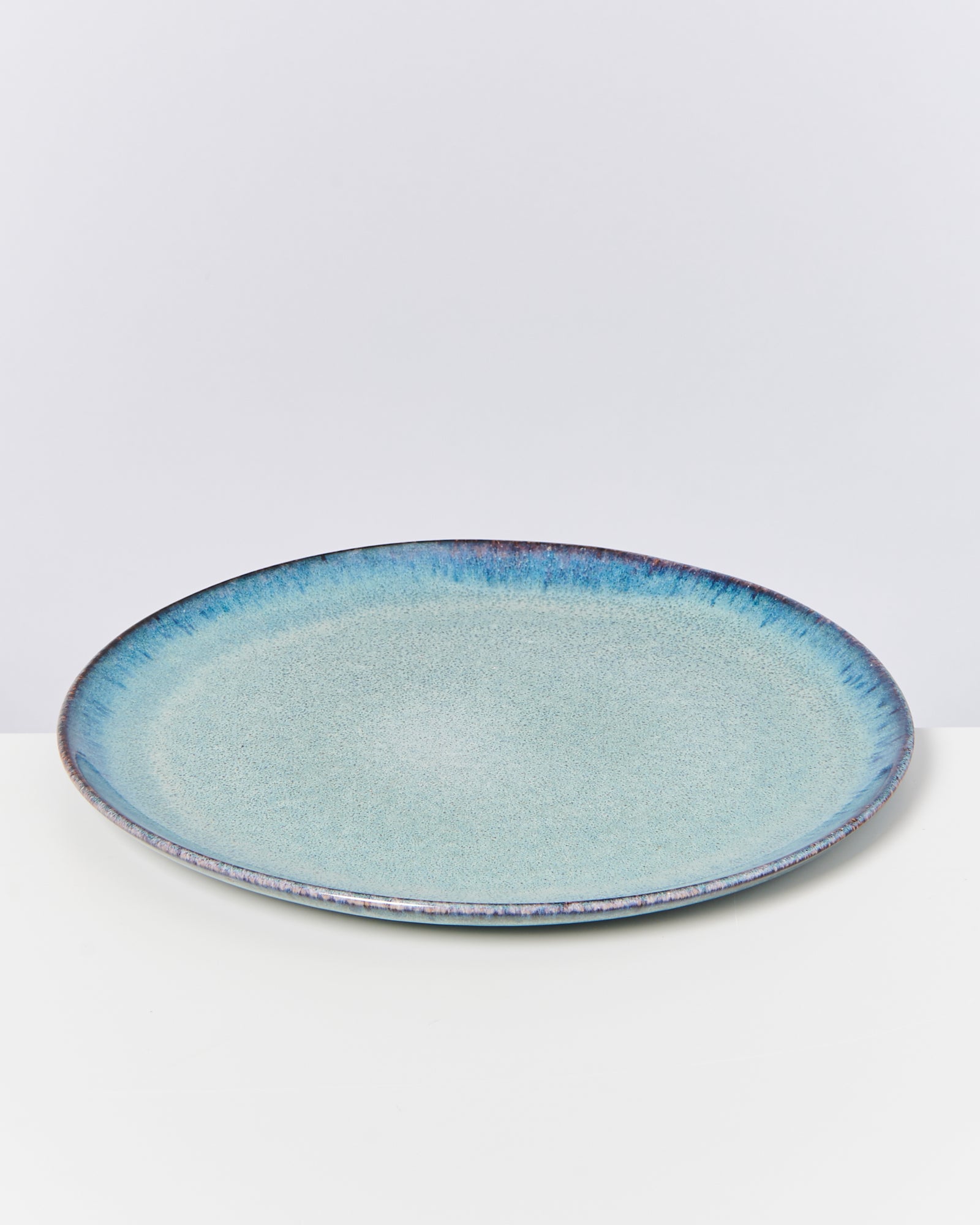 Großer, runder, flacher Teller mit blauer und leicht grünlicher Glasur auf weißem Hintergrund.