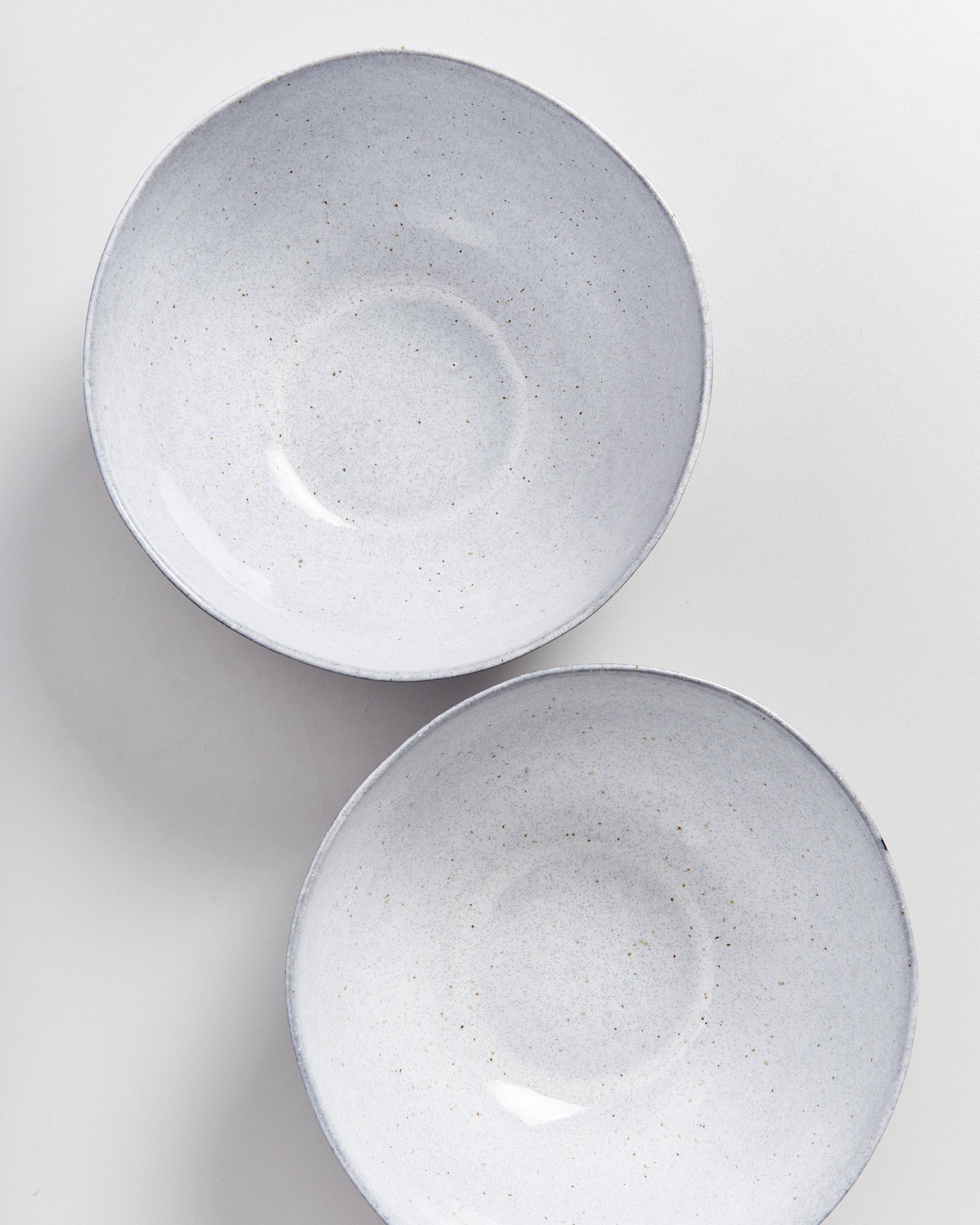 Zwei helle Keramikschalen mit feinen Sprenkeln auf einer weißen Oberfläche, von oben betrachtet.