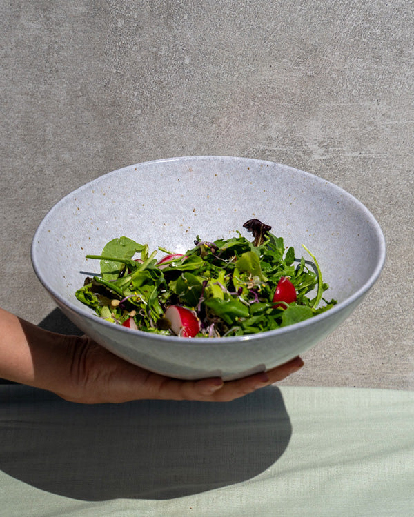 Hand hält eine Schüssel mit gemischtem grünen Salat und Radieschen vor grauem Hintergrund.
