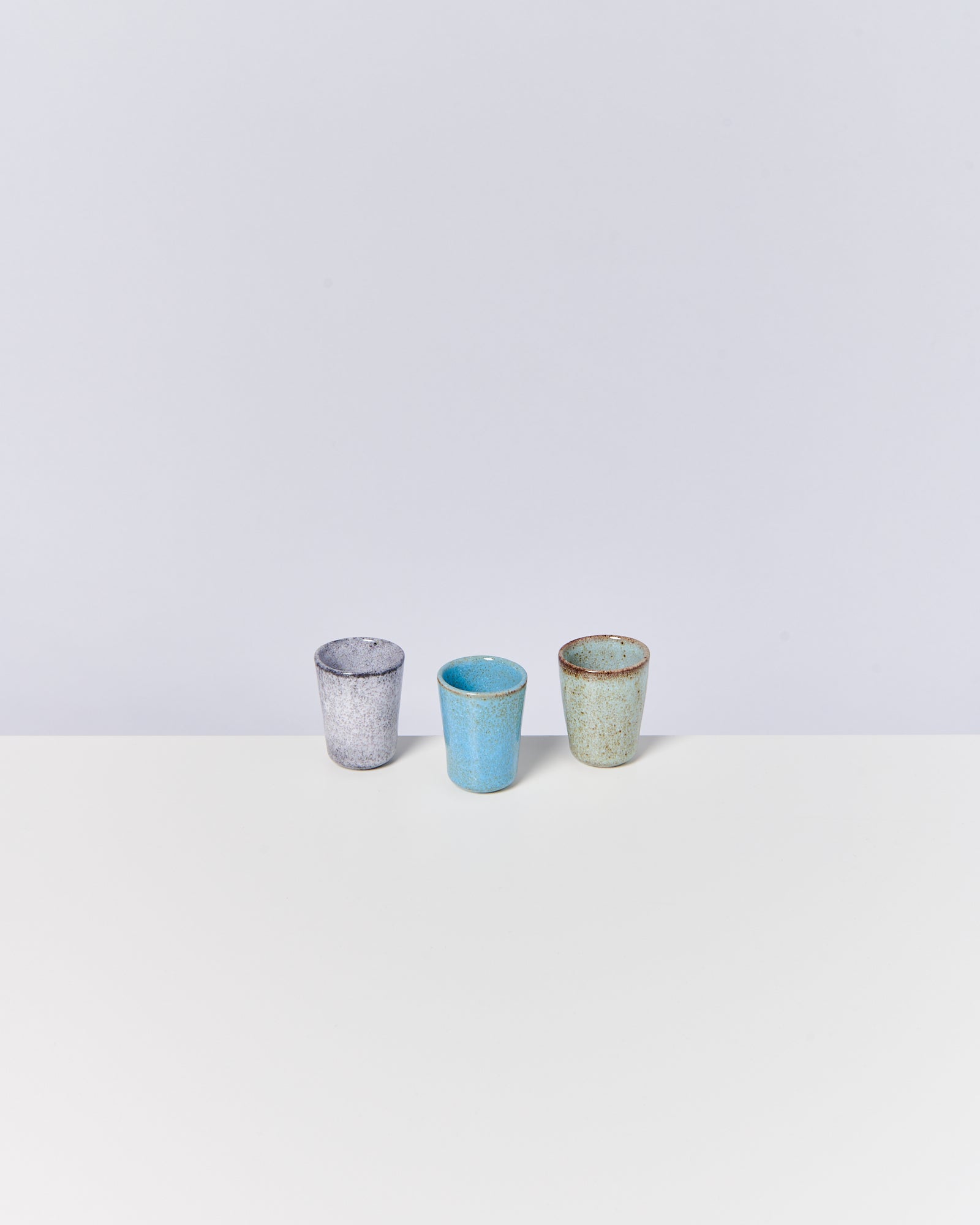 Drei bunte, keramikähnliche Becher auf einer weißen Fläche vor grauem Hintergrund.