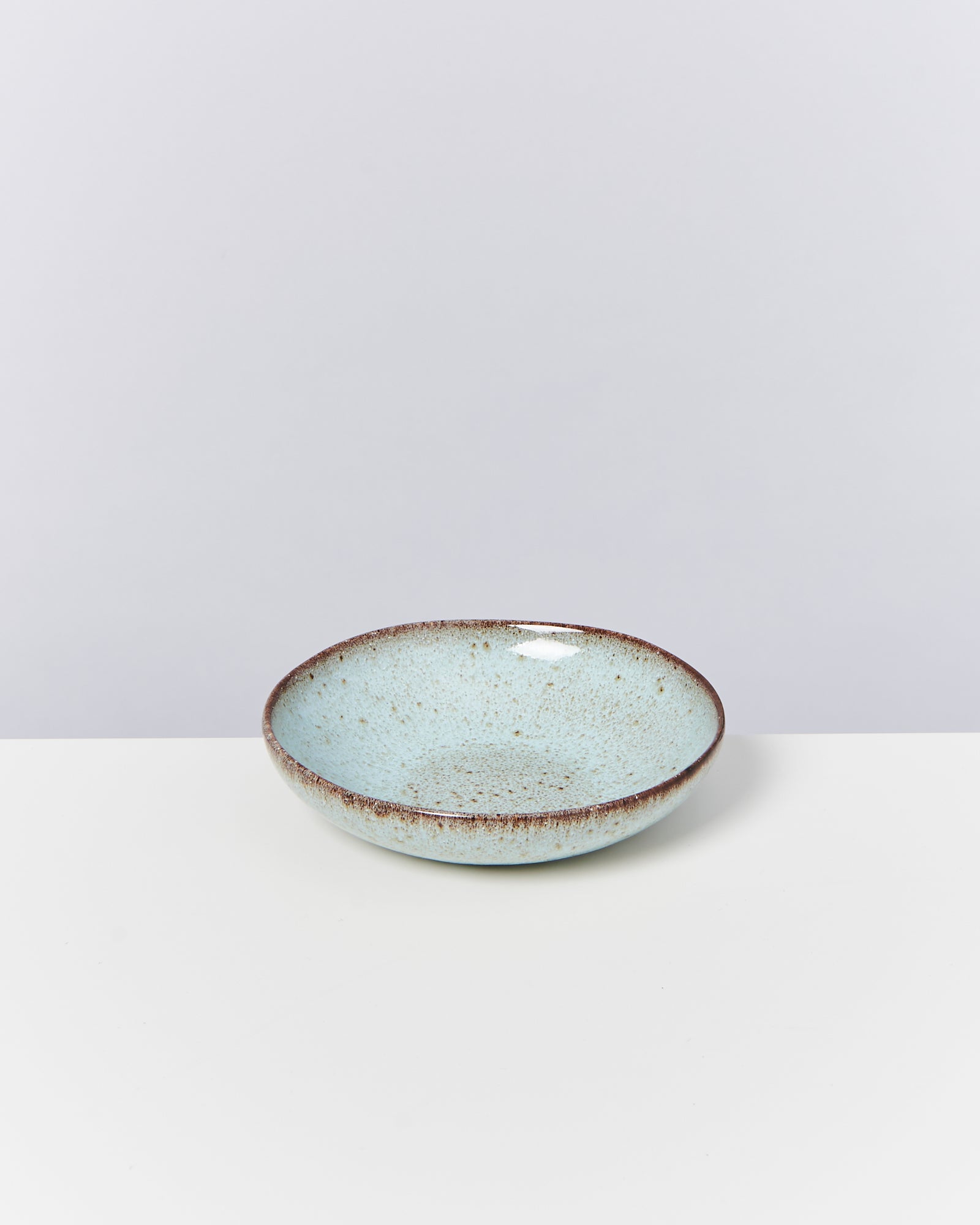 Eine hellblaue, gesprenkelte Keramikschüssel mit braunem Rand steht auf einem weißen Tisch vor grauem Hintergrund.