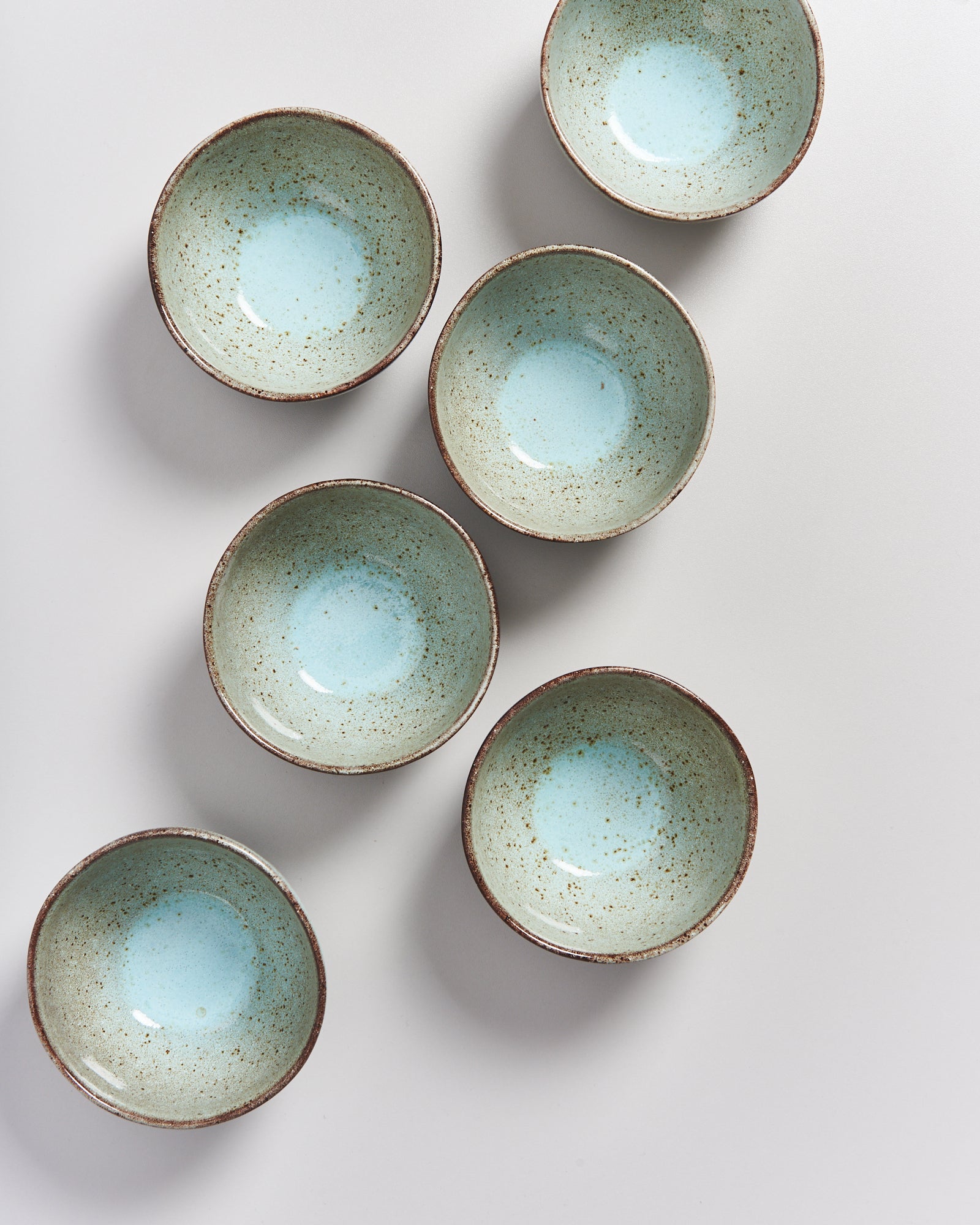 Sechs türkisfarbene Keramikschüsseln mit braunem Rand sind auf einer weißen Oberfläche angeordnet.
