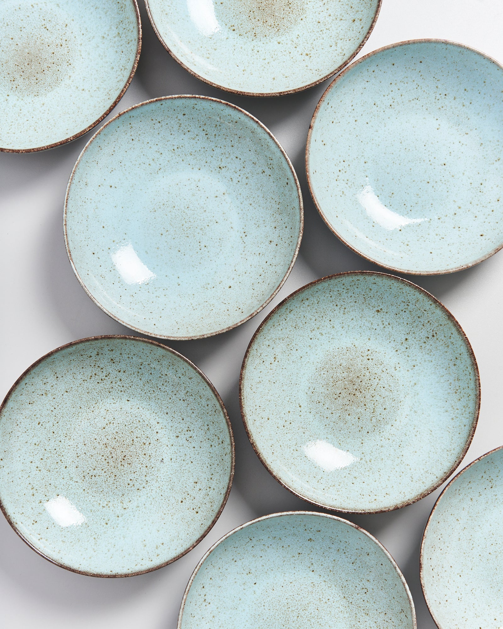 Acht hellblaue, gesprenkelte Keramikteller mit braunen Rändern liegen auf einer Fläche verteilt.