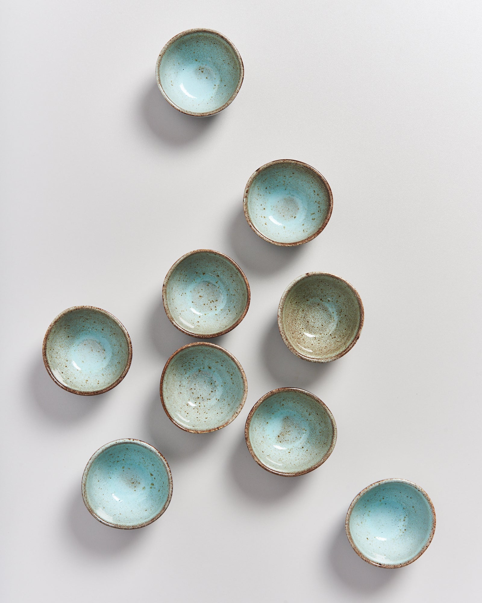 Neun kleine, bläulich glasierte Schalen mit braunem Rand auf einer grauen Fläche.
