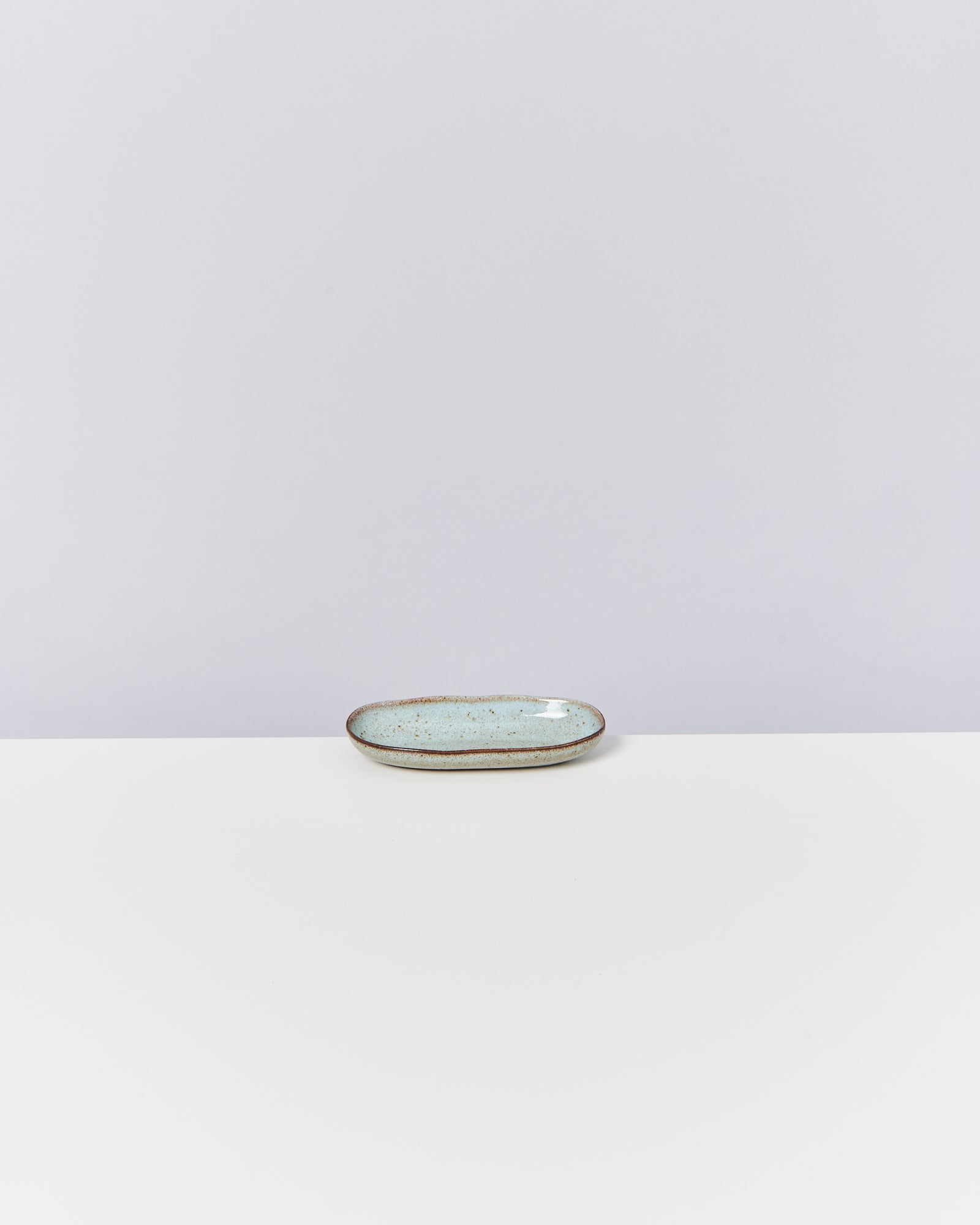 Kleiner, ovale, hellblauer Keramikteller mit braunem Rand auf weißem Untergrund vor grauem Hintergrund.