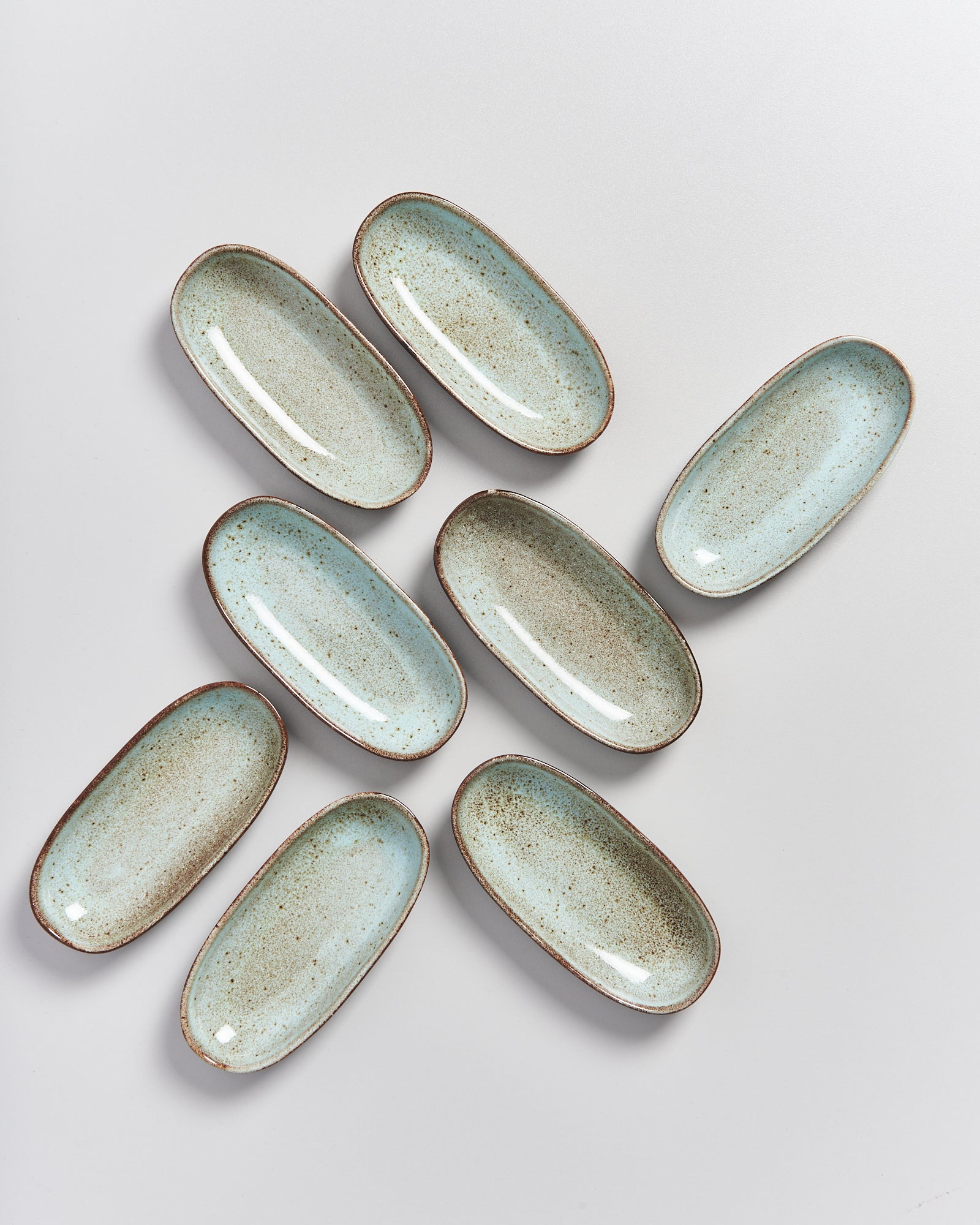 Acht längliche Keramikteller mit blauer Glasur und braunem Rand liegen auf einem weißen Hintergrund.