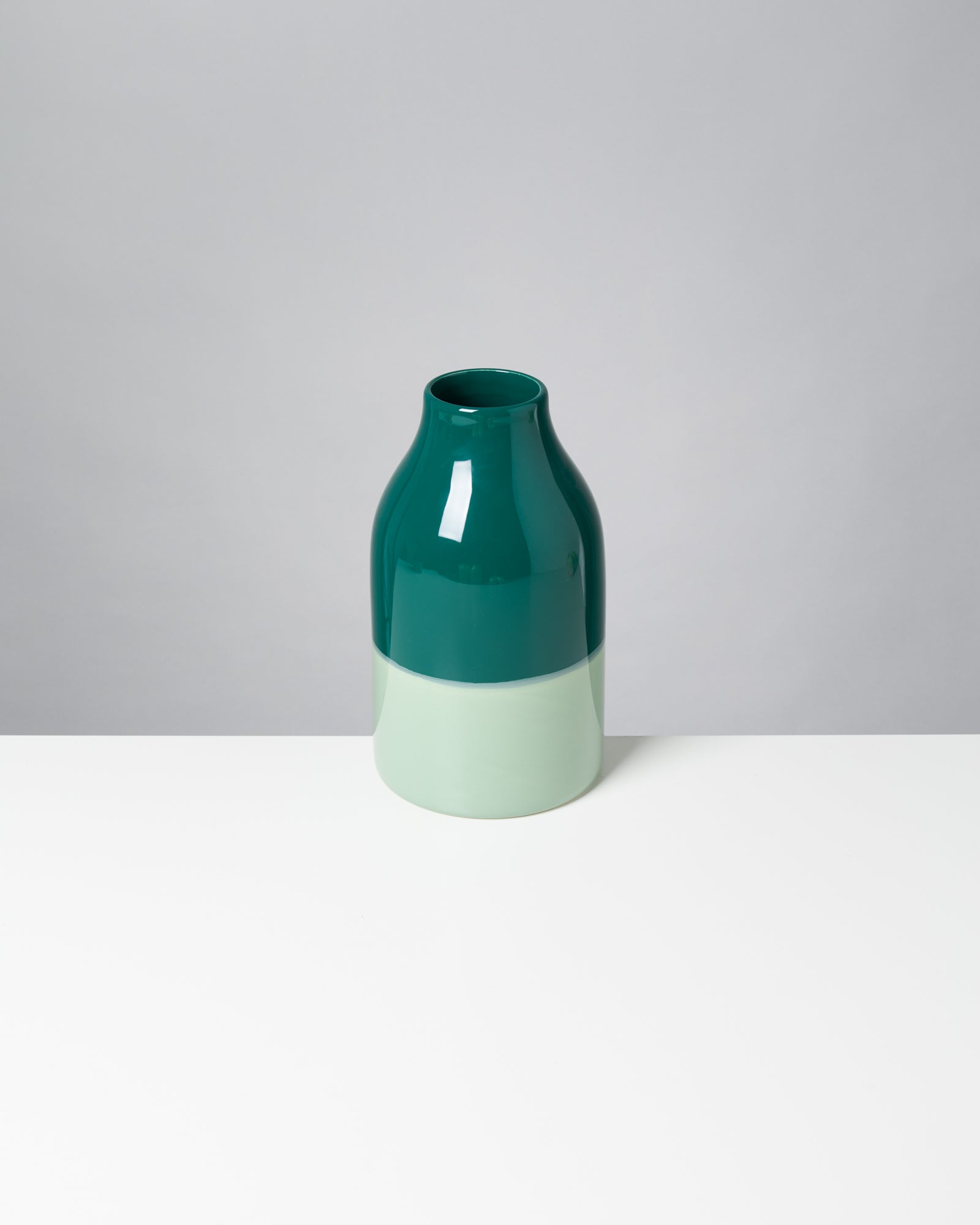 Grüne Vase mit glänzender Oberfläche steht auf einem weißen Tisch vor grauem Hintergrund.