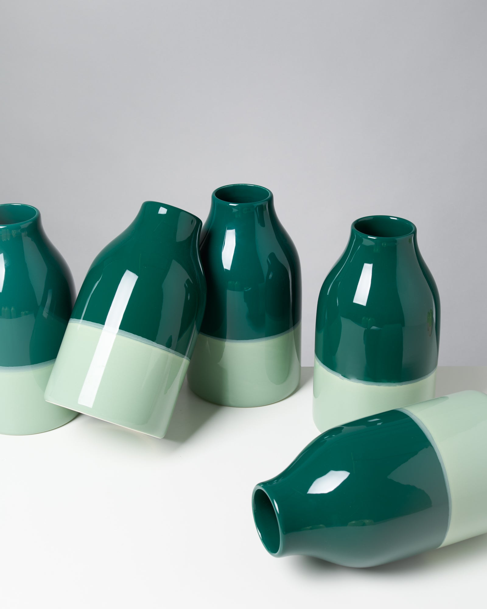 Fünf grün-weiße, glänzende Keramikvasen stehen und liegen auf einer hellen Oberfläche.