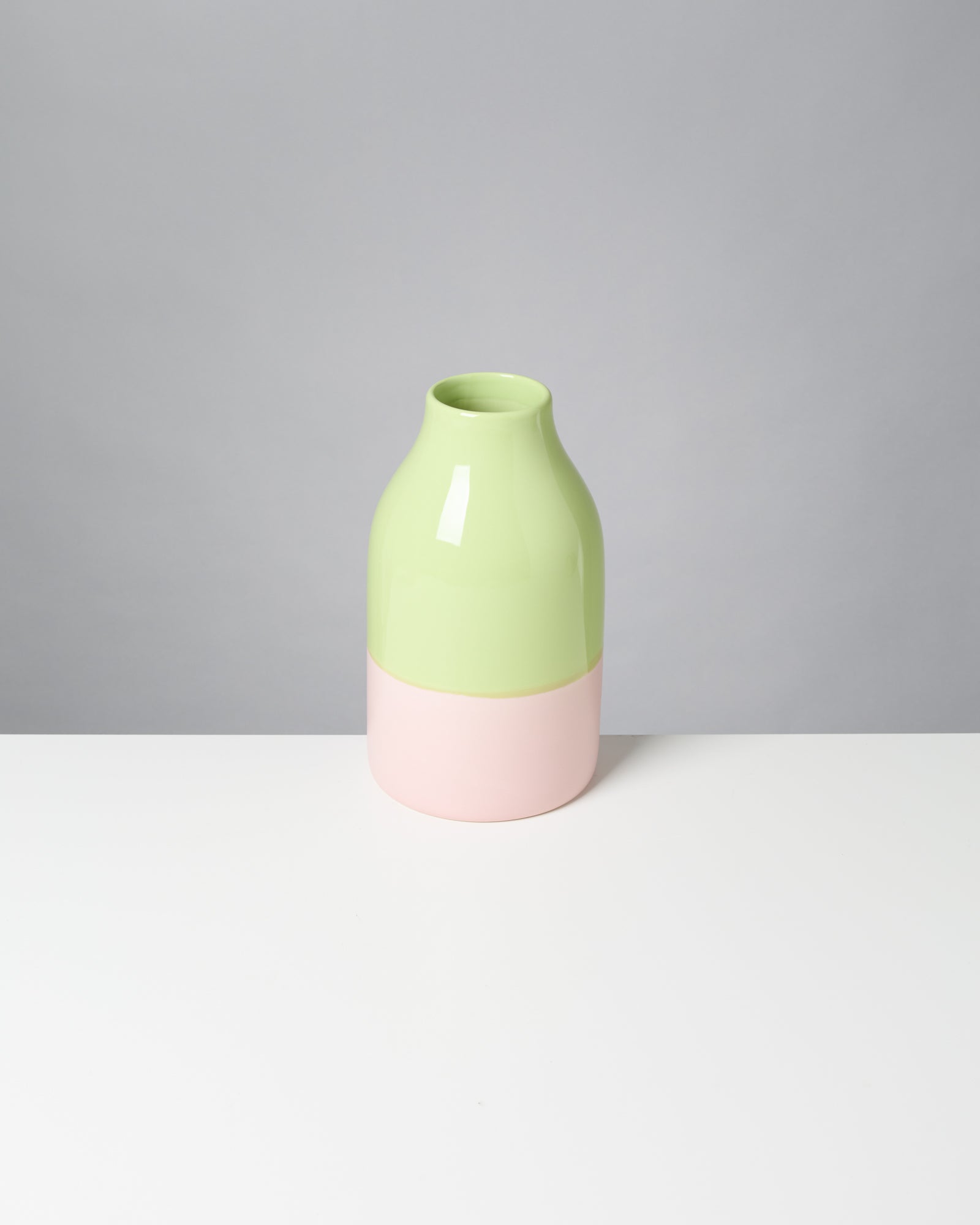 Eine zweifarbige Vase, oben hellgrün und unten rosa, steht auf einer weißen Oberfläche vor einem grauen Hintergrund.