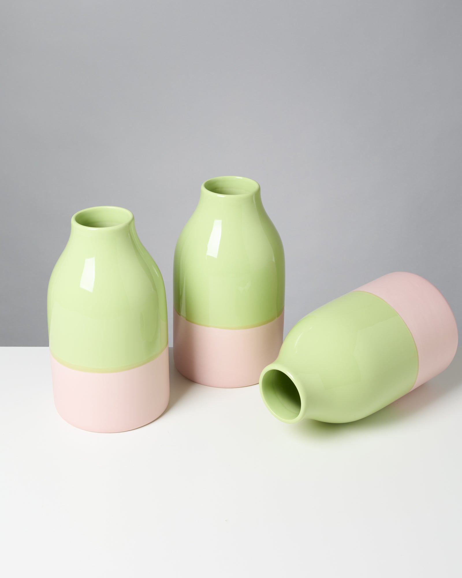 Drei zweifarbige Vasen in Grün und Rosa auf einem weißen Tisch, eine Vase liegt seitlich.