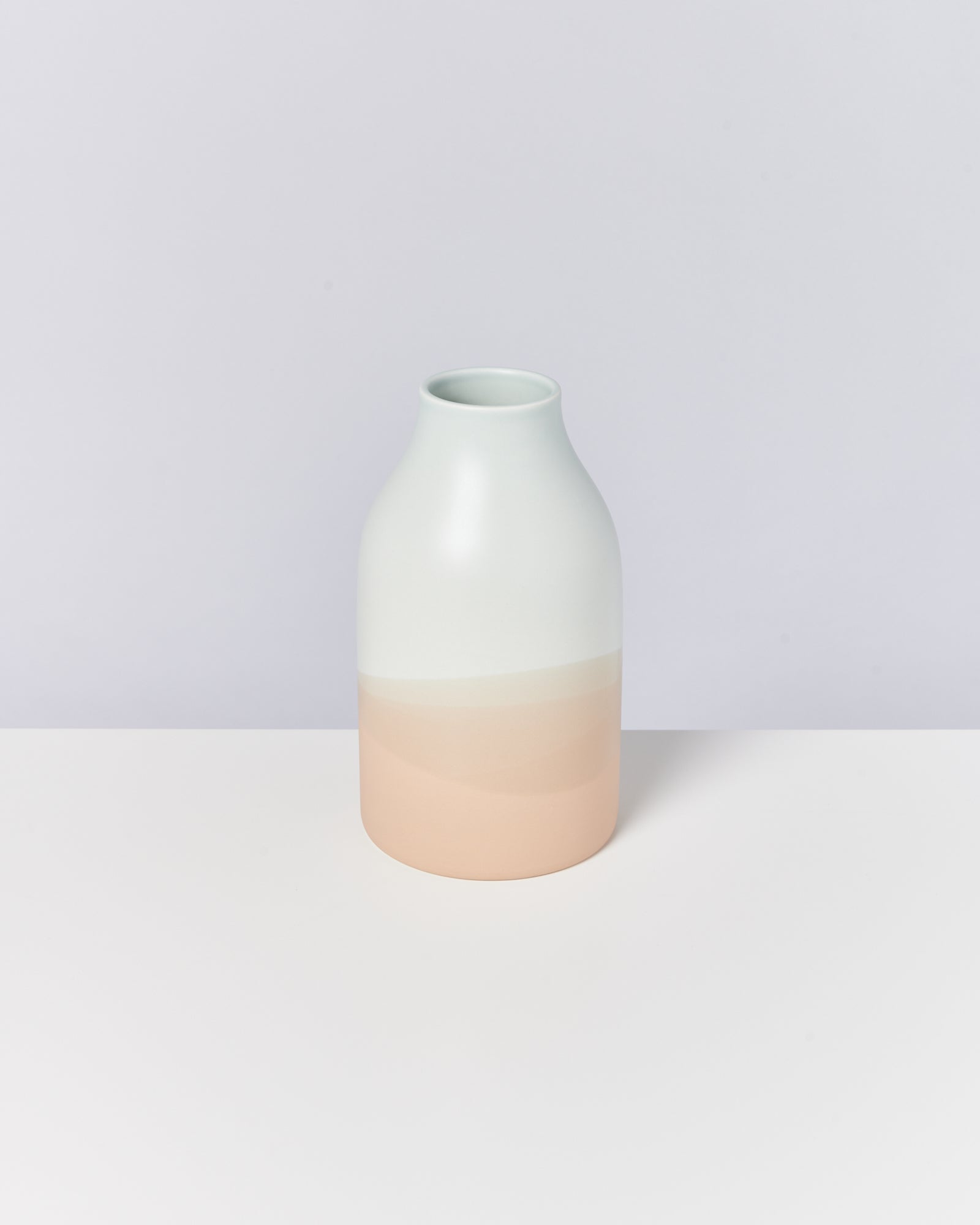 Minimalistische Vase in mattem Farbverlauf von Weiß nach Hellrosa, auf einem hellen Hintergrund stehend.