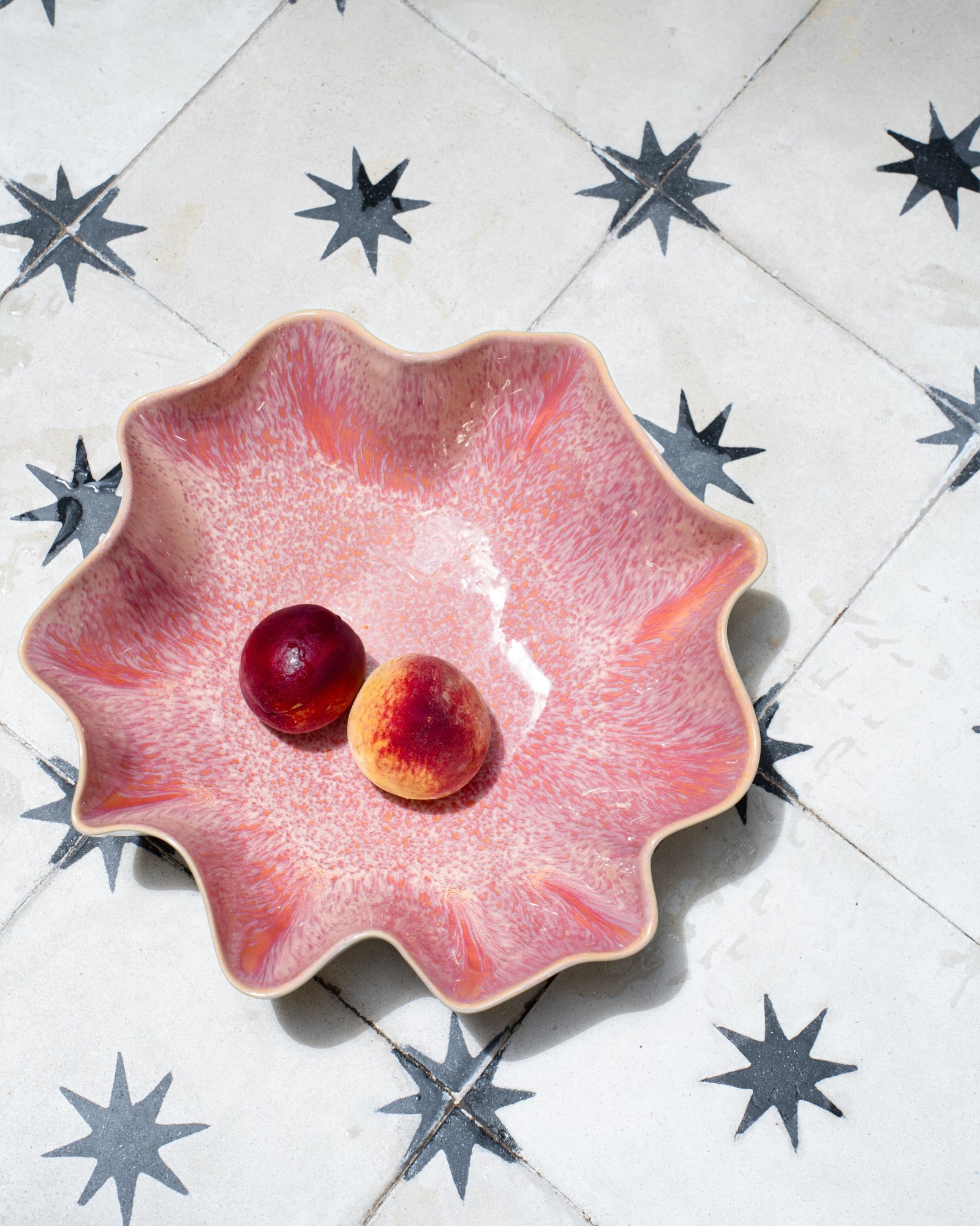 Zwei Pfirsiche liegen in einer rosafarbenen, wellenförmigen Schale auf einem mit sternförmigen Fliesen bedeckten Boden.