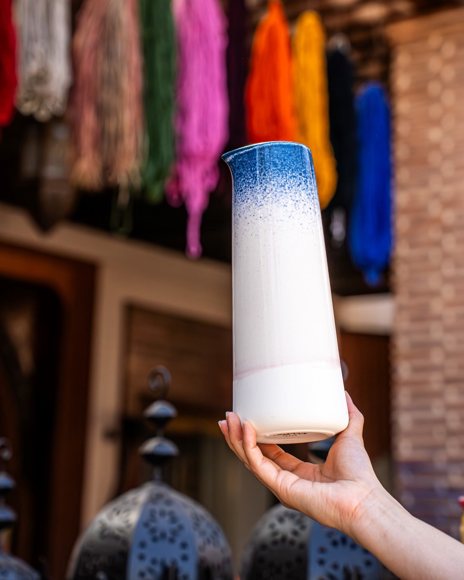 Person hält eine hohe Keramikvase mit blau-weißem Verlauf, Hintergrund mit bunten Fäden und traditionellen Laternen.