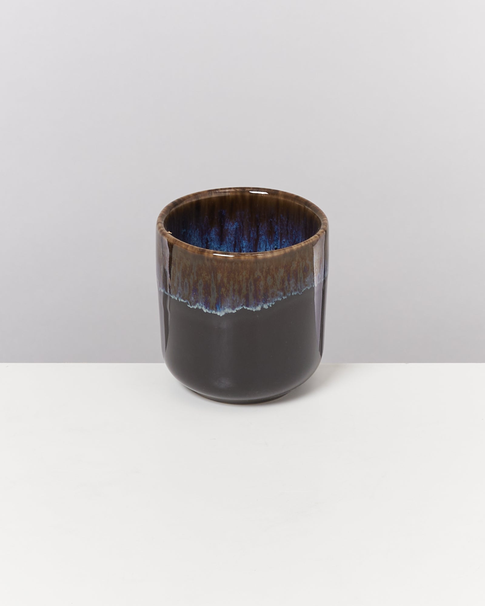 Dunkler, glasanmutender Becher mit blaublauer Innenflächenglasur auf einem weißen Tisch vor grauem Hintergrund.
