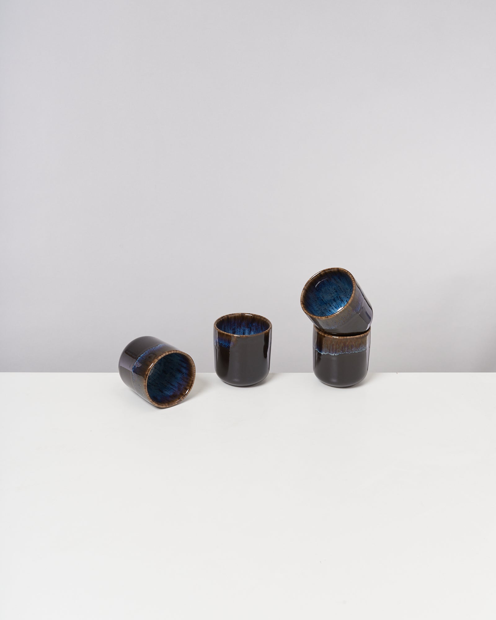 Vier schwarze Keramiktassen mit blauer Innenseite auf einem weißen Tisch vor grauem Hintergrund.