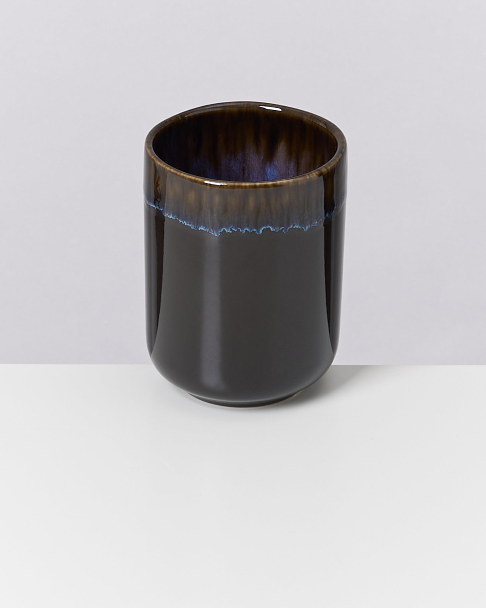 Schwarzer, glänzender Keramikbecher mit blauer Linie am oberen Rand auf einem weißen Tisch vor grauem Hintergrund.