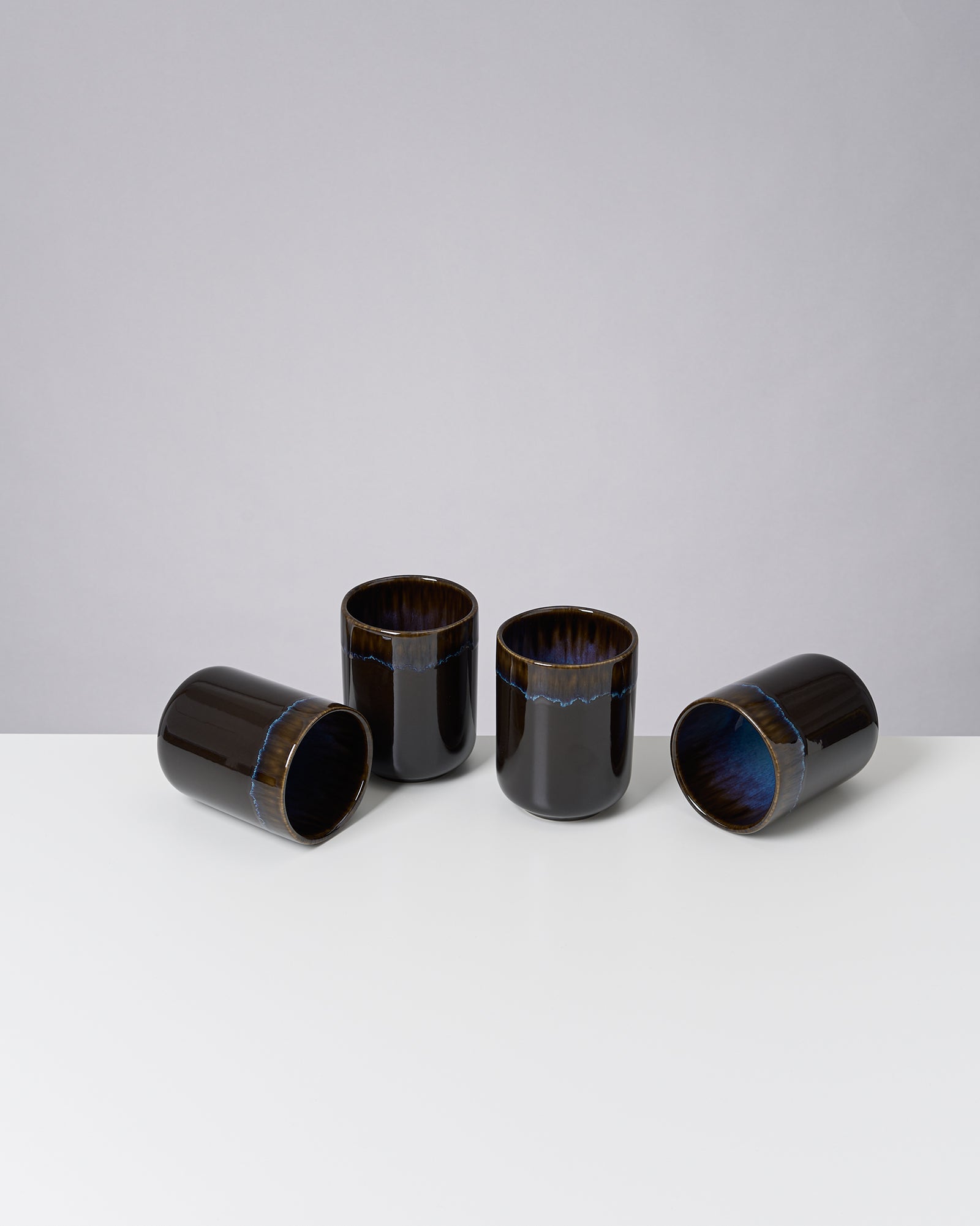 Vier dunkle Keramikbecher, drei aufrecht und einer liegend, auf einem weißen Tisch vor grauem Hintergrund.