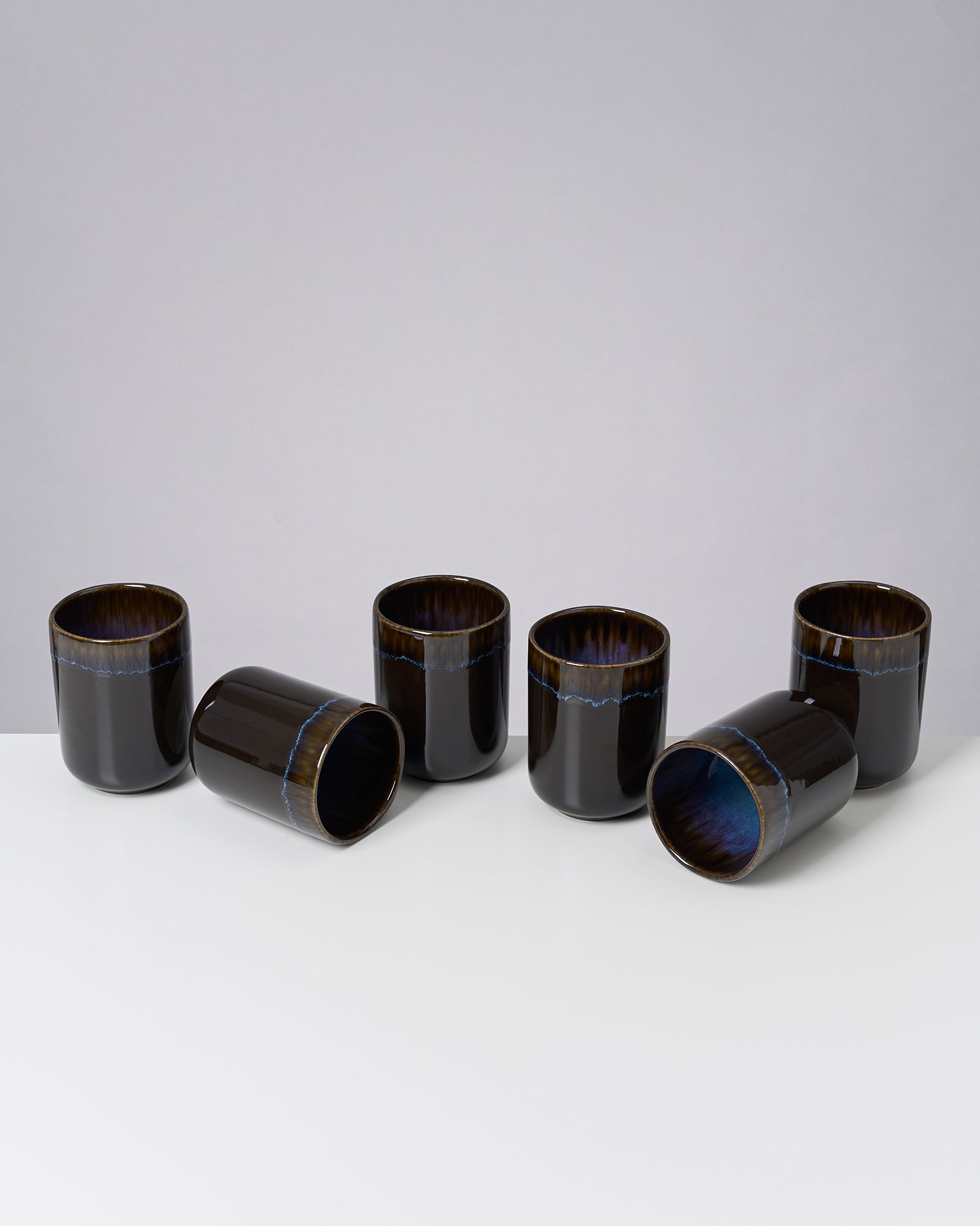 Sechs schwarze Becher mit blauen Akzenten, zwei liegen seitlich, auf einer weißen Oberfläche vor grauem Hintergrund.