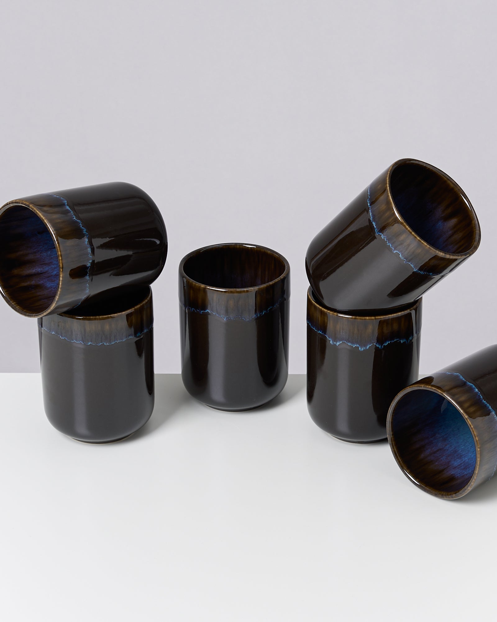 Sechs dunkelblaue, glänzende Becher stehen und liegen auf einem weißen Tisch vor grauem Hintergrund.