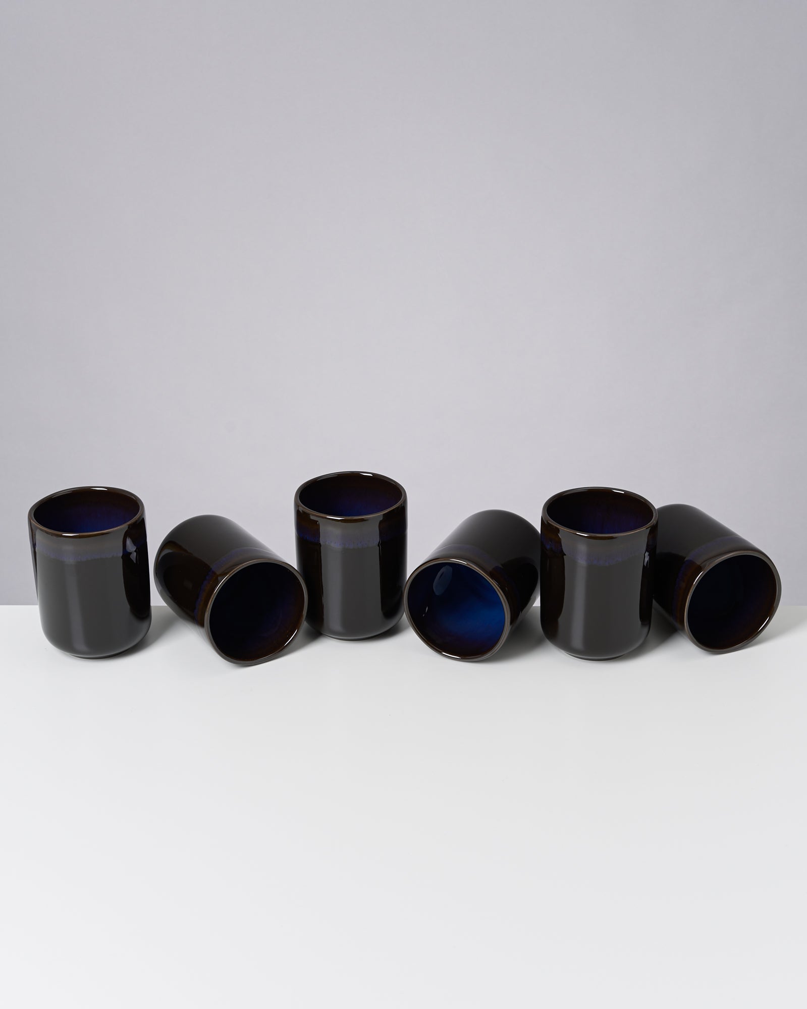Sechs schwarze Tassen, teilweise liegend, auf einem weißen Tisch vor grauem Hintergrund.