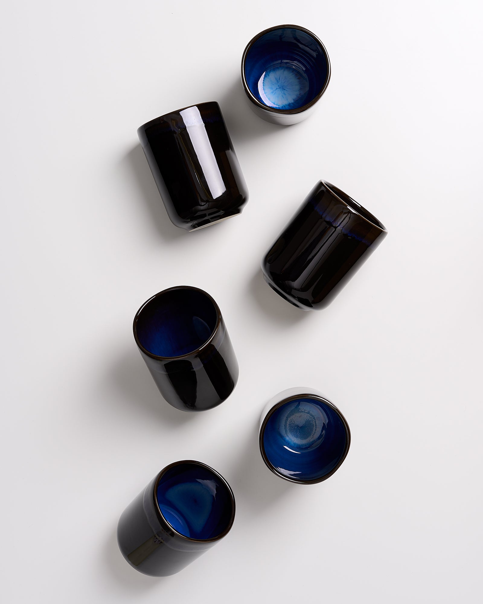 Sechs schwarze Becher mit blauem Inneren, unregelmäßig auf weißem Hintergrund verteilt.
