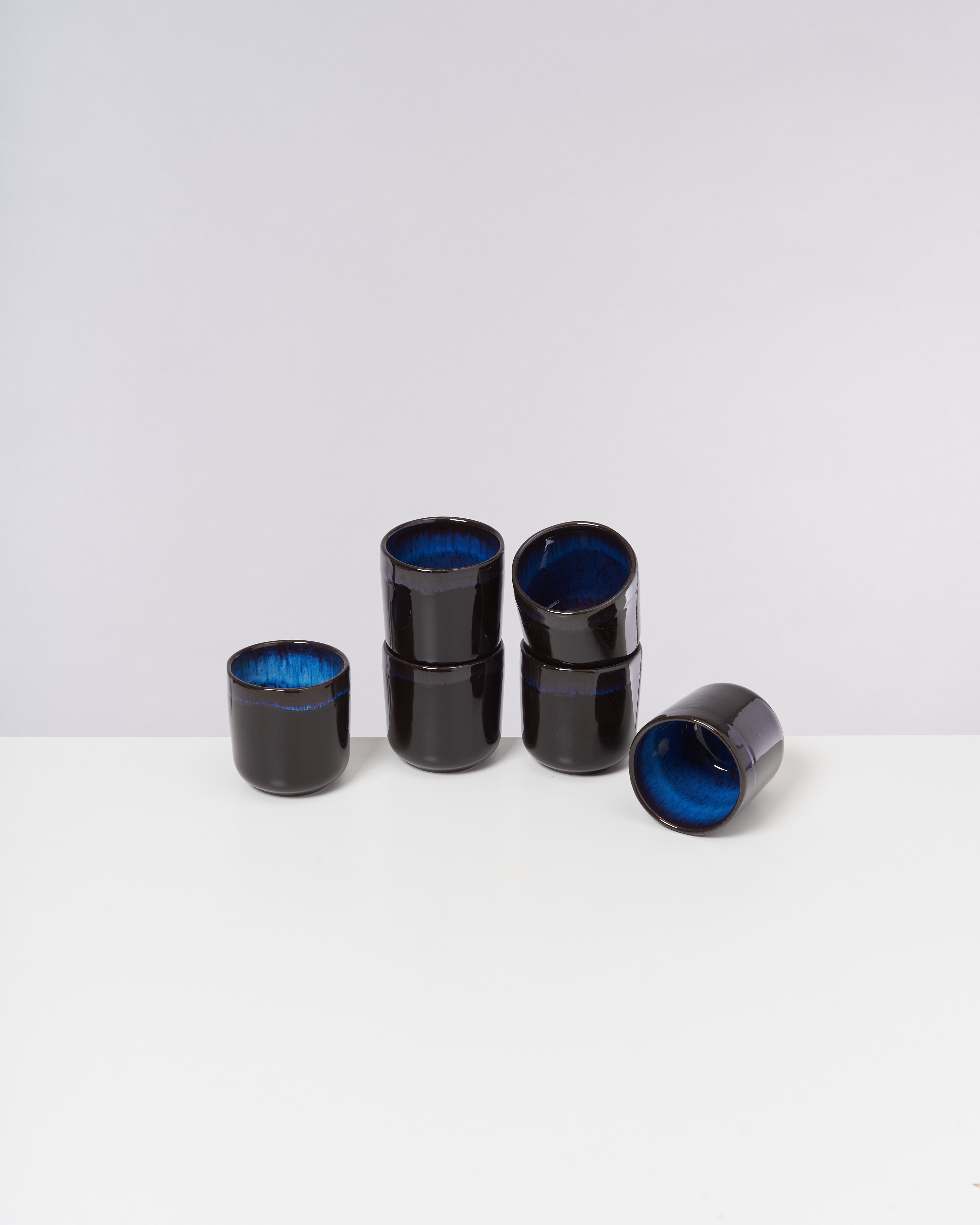 Fünf schwarze Keramiktassen mit blauer Innenseite stehen auf einem weißen Tisch vor einem grauen Hintergrund.