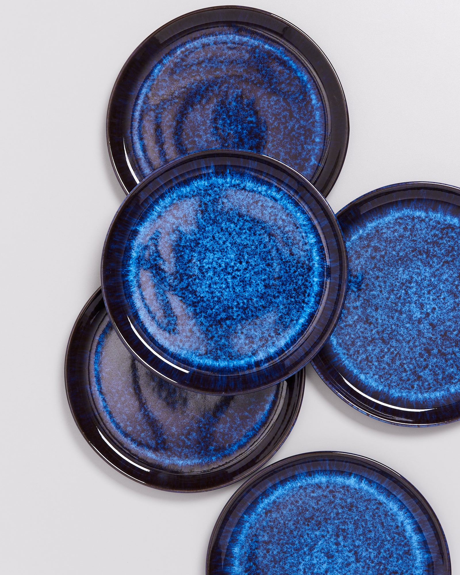 Fünf blaue Keramikteller mit dunklen Rand und marmorierten Mustern liegen überlappend auf einer hellgrauen Oberfläche.