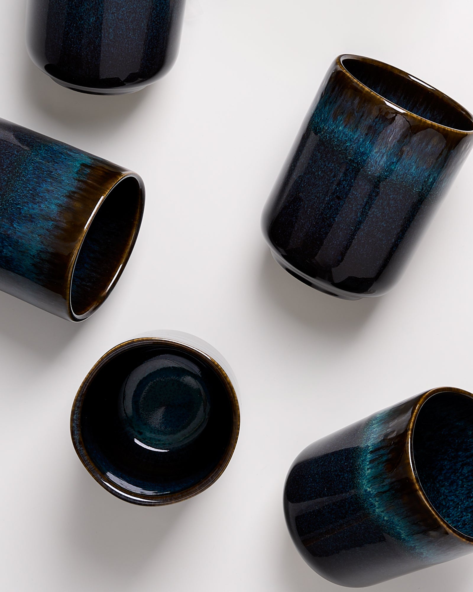 Fünf schwarze Becher mit blauer und brauner Glasur liegen auf einem weißen Hintergrund.