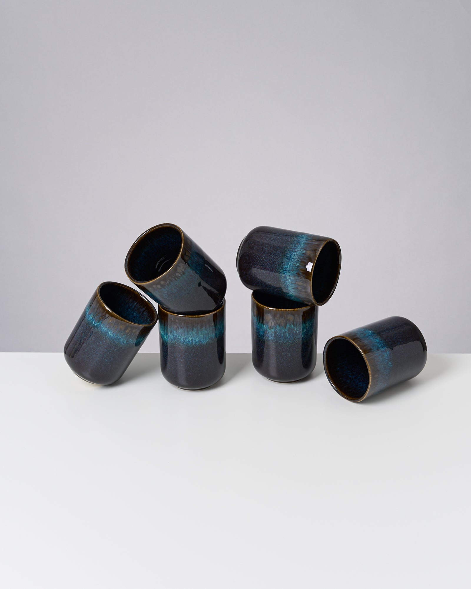 Sechs dunkelblaue Keramikbecher, drei gerade und drei umgekippt, auf einer weißen Oberfläche vor grauem Hintergrund.
