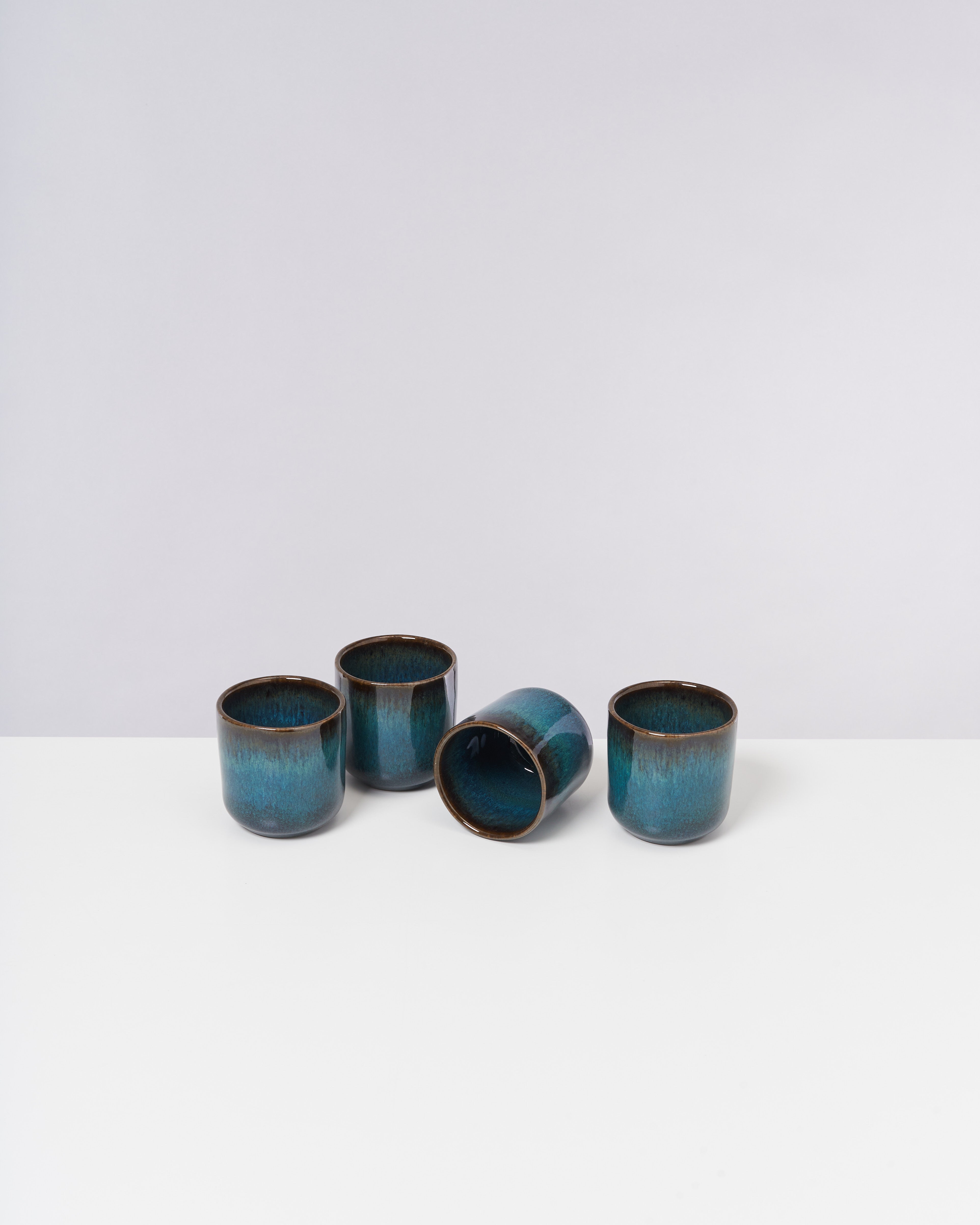 Vier blaue Keramiktassen, eine davon umgekippt, auf einem weißen Tisch vor einer grauen Wand.