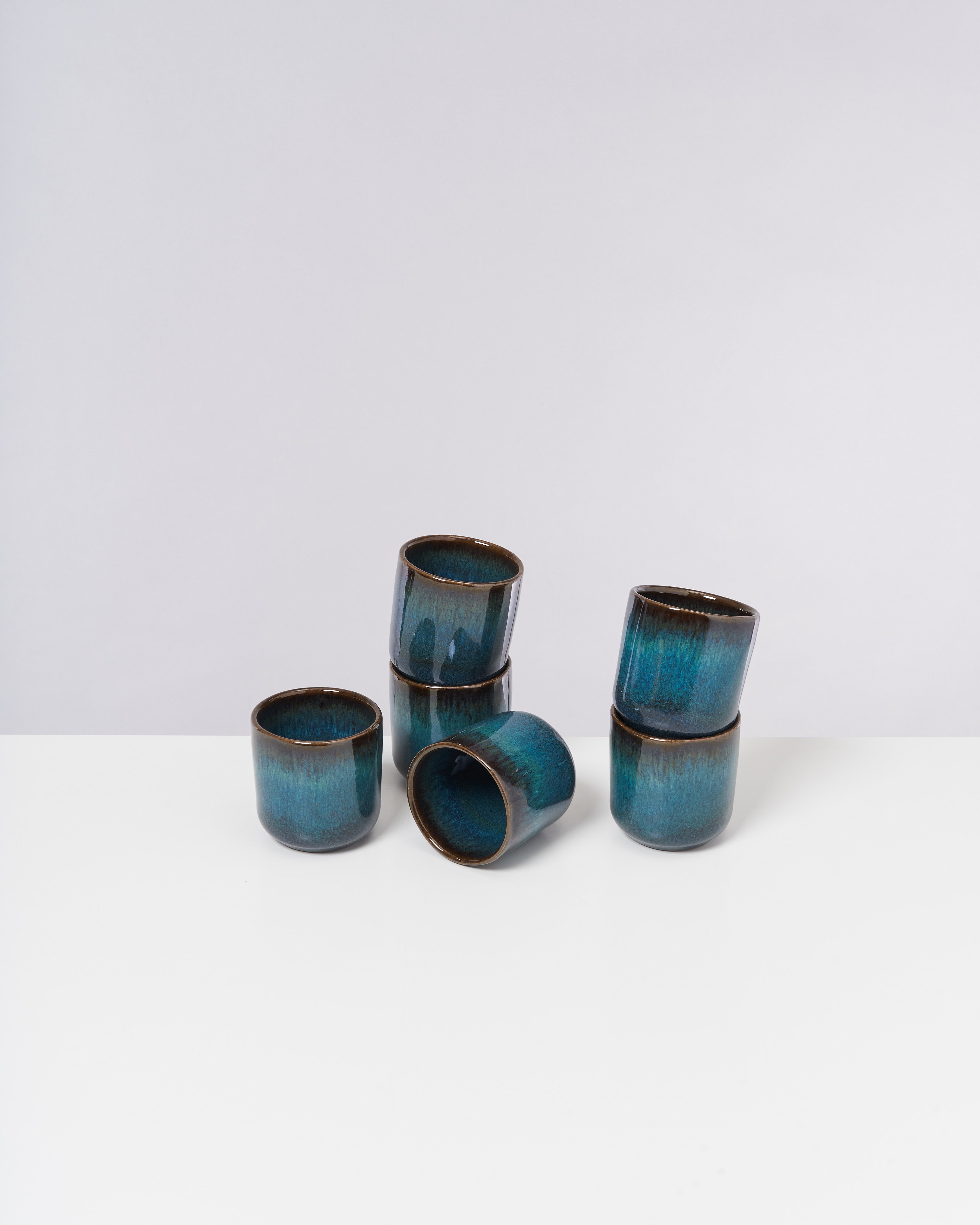 Sechs blaue, gläserne Becher stehen auf weißem Hintergrund, einige sind gestapelt.