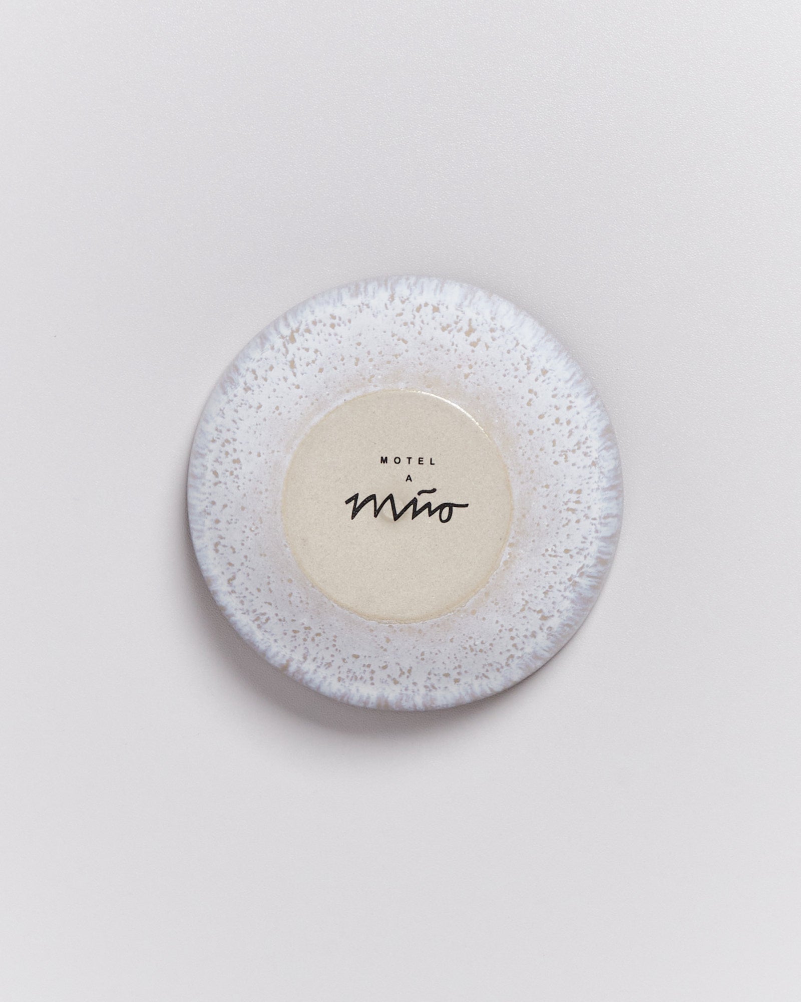 Weiße Keramikschale mit hellbraunem Kreis in der Mitte und der Aufschrift "Motel a Miio" auf einer grauen Oberfläche.