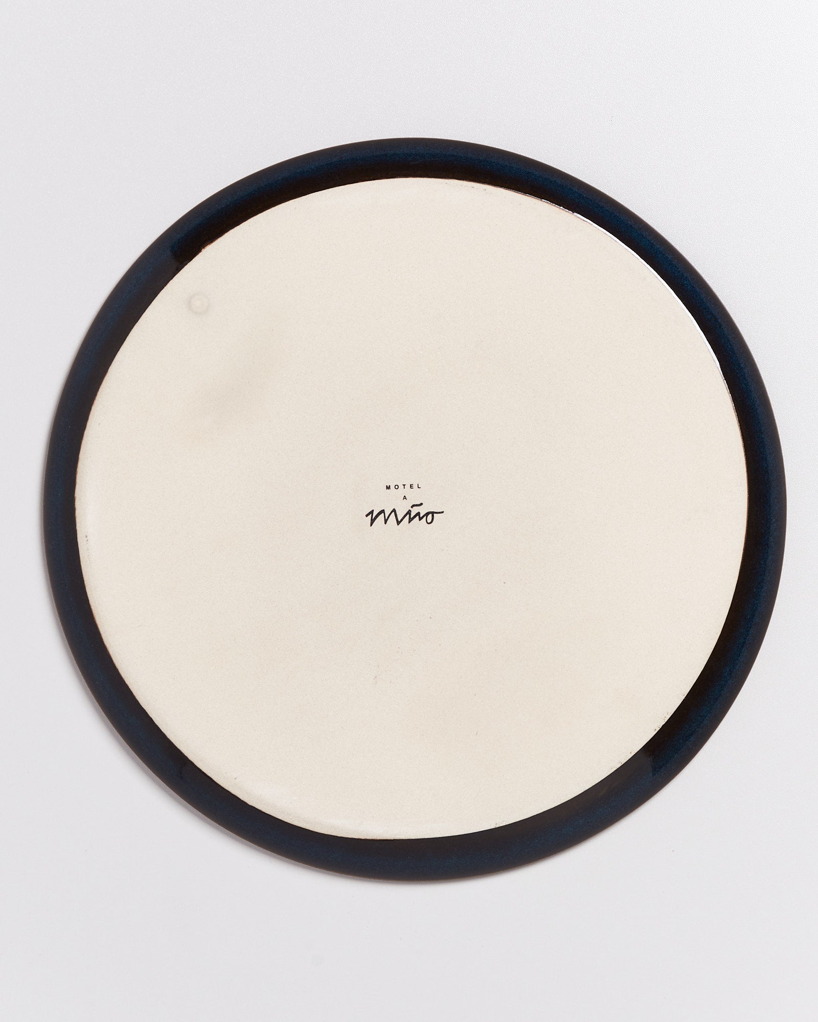 Rückseite eines weißen Tellers mit einem dunklen Rand und einer kleinen, mittigen Markierung oder Unterschrift.