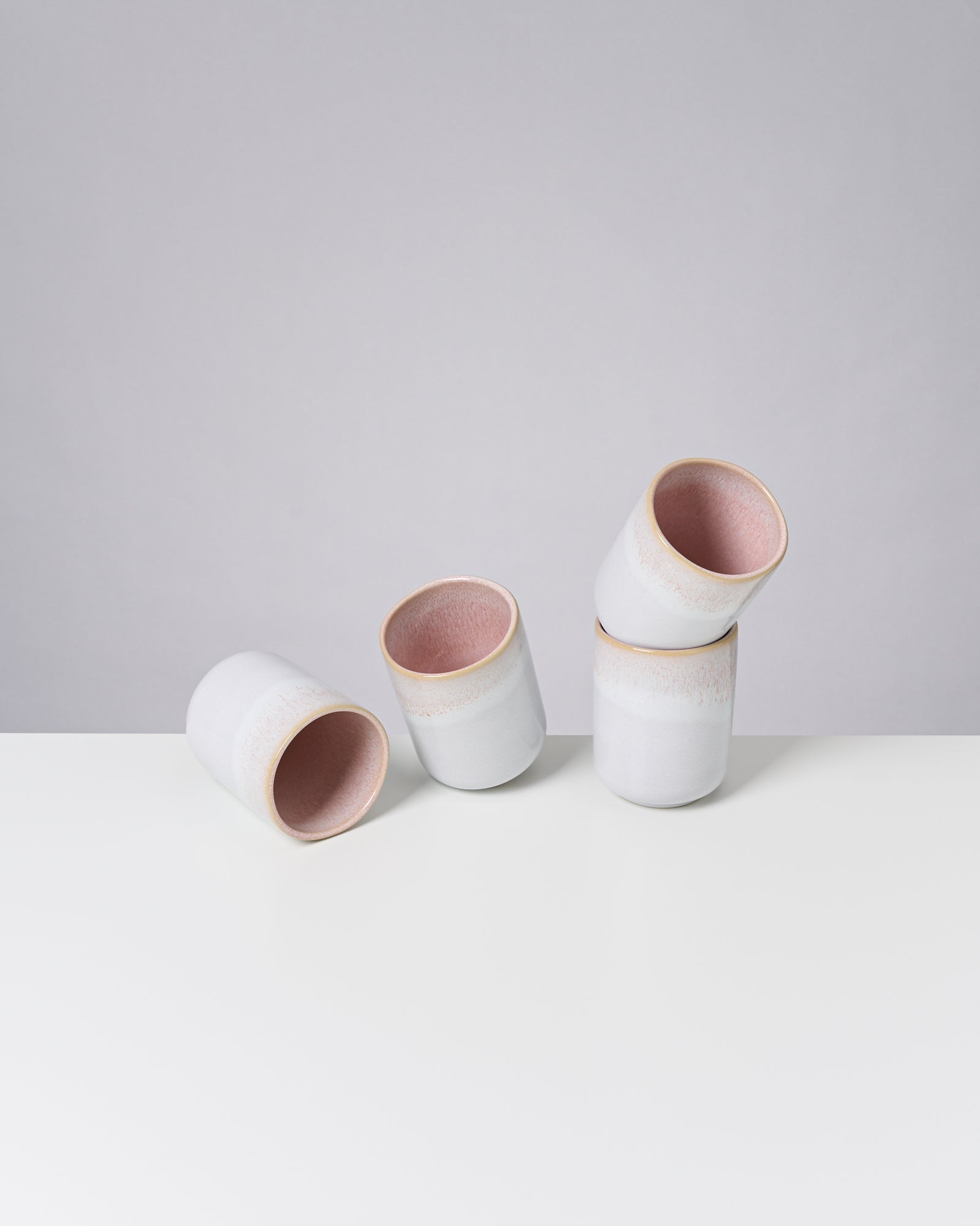 Vier weiße Keramikbecher mit rosa Innenflächen stehen und liegen auf einem weißen Tisch.