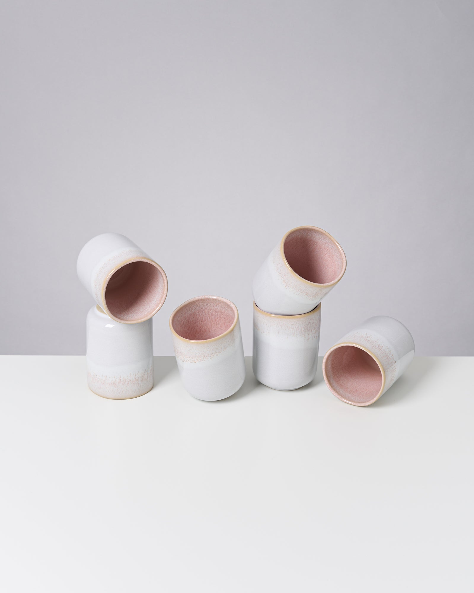 Sechs weiße Keramiktassen mit rosa Innenraum, ungleichmäßig auf einem weißen Tisch arrangiert.