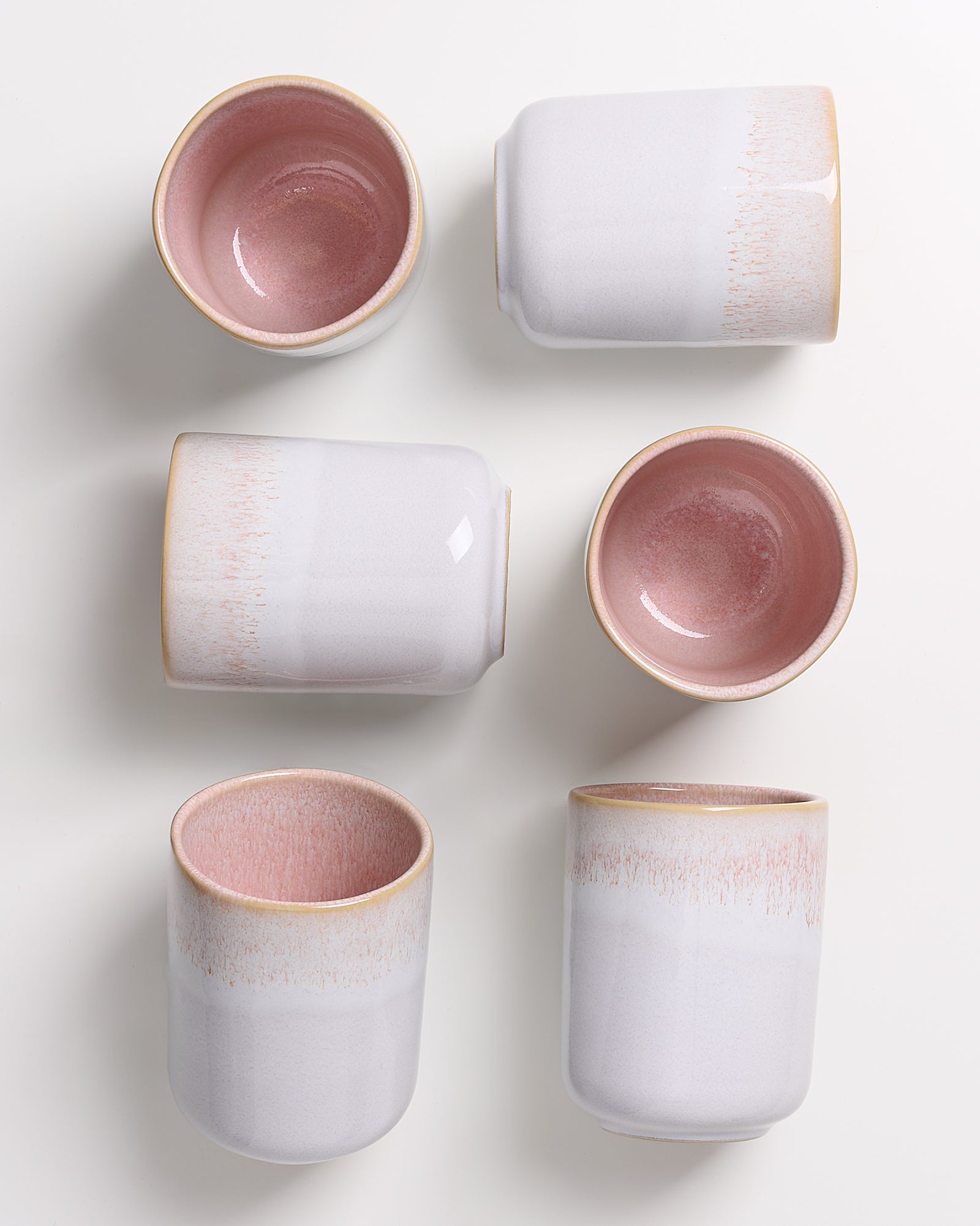 Sechs Keramikbecher in Rosa und Weiß auf weißem Hintergrund, teilweise liegend, teilweise stehend.